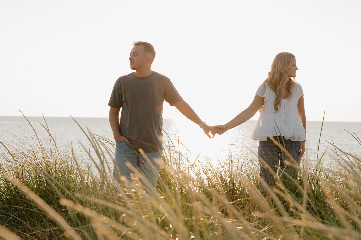 Silhouetted couple walking through tall beach grass during golden hour by the water.
