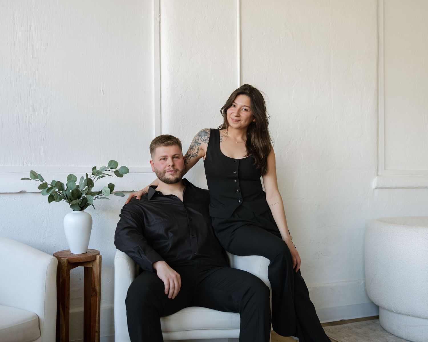 A man and woman in black outfits pose elegantly in a bright white room with a vase of greenery nearby.