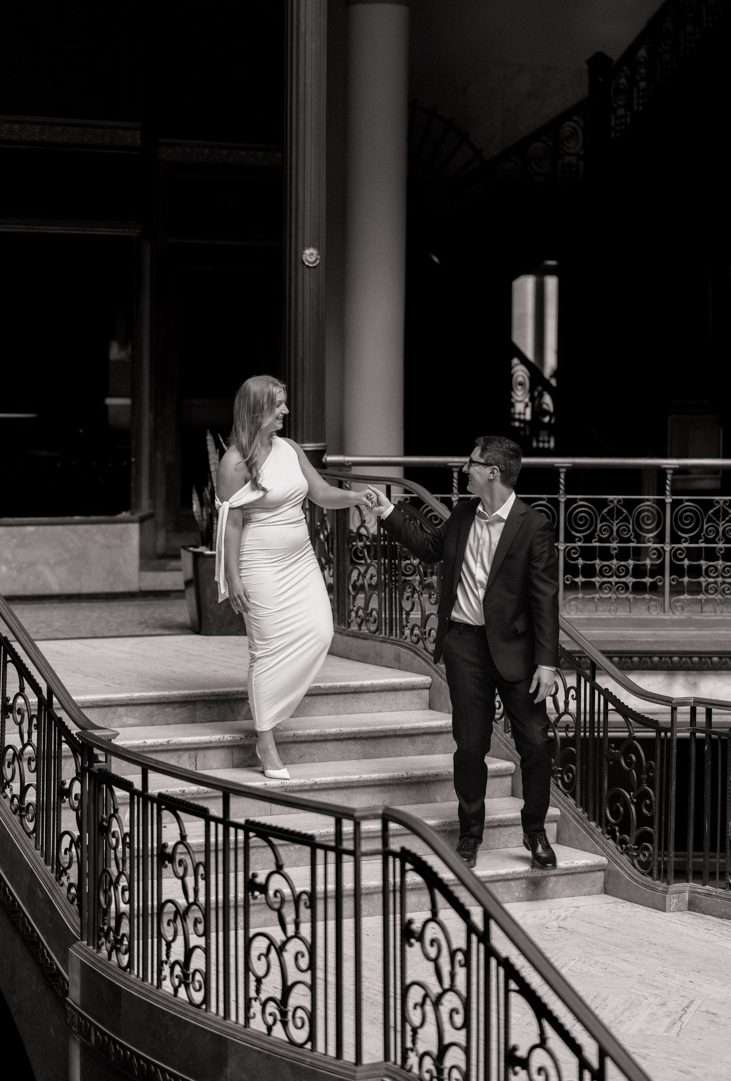 Black and white photo of a couple holding hands on elegant staircase, woman in white dress, man in dark suit.