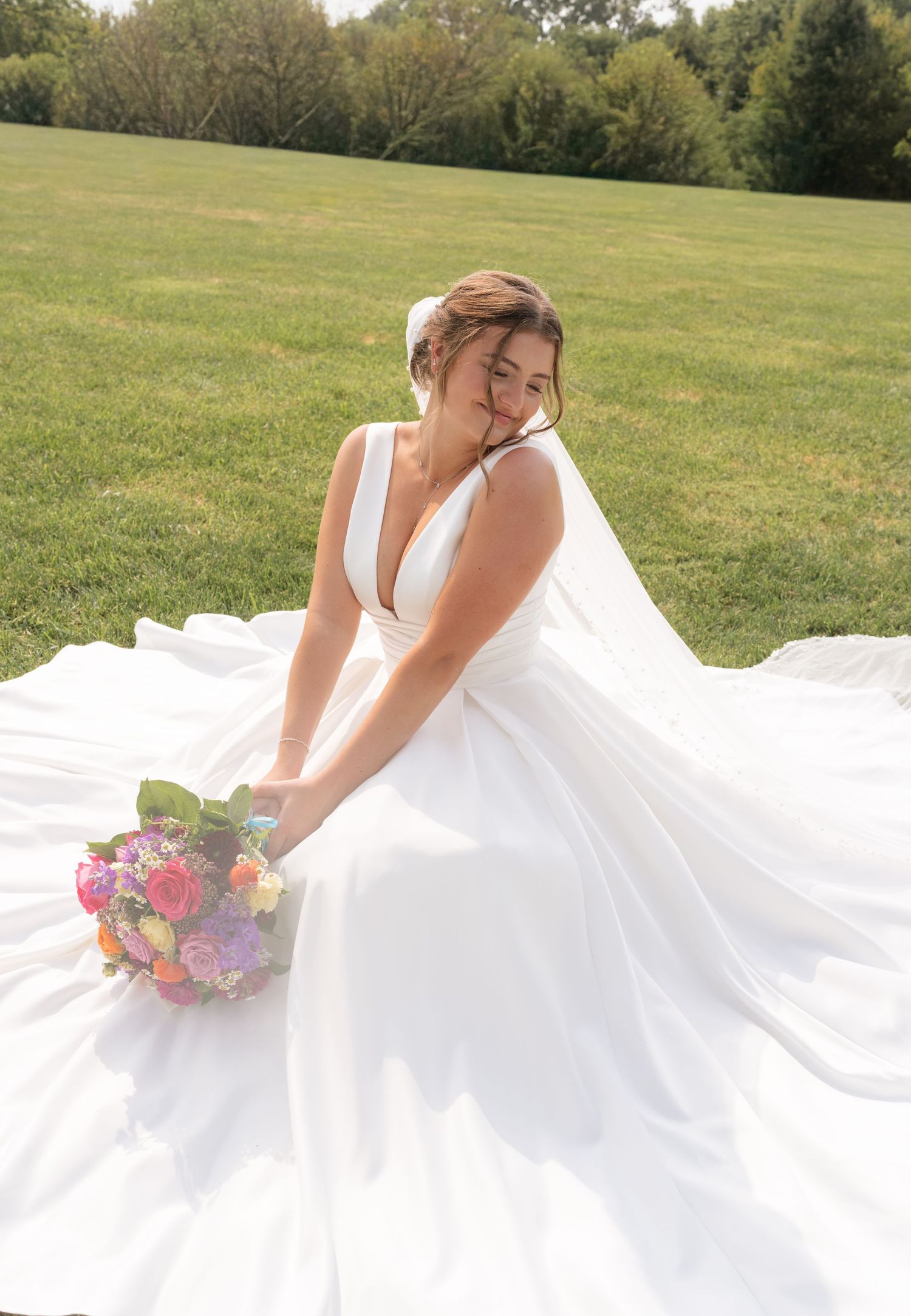 Bride in a white ball gown with deep V-neck sits on green lawn holding a colorful bouquet, smiling over her shoulder.