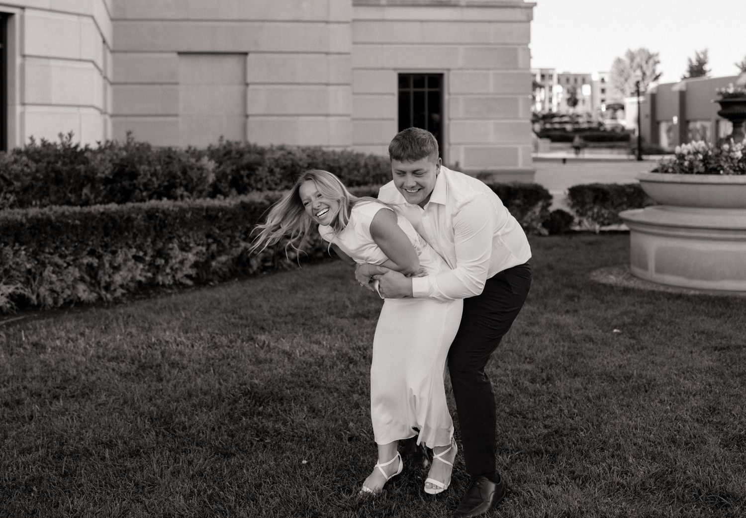 Black and white photo of a couple dancing outdoors, man dipping woman in white dress, laughing joyfully near a building.