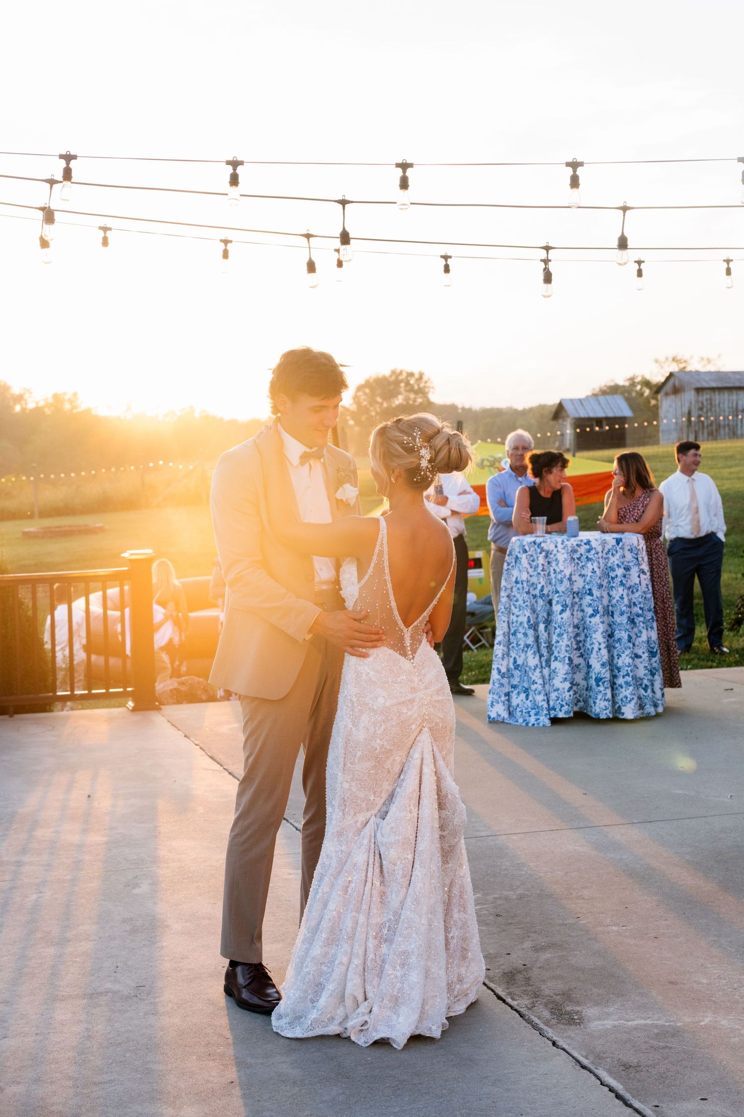 Bride and groom share first dance outdoors at golden hour, with string lights overhead and guests mingling in background.