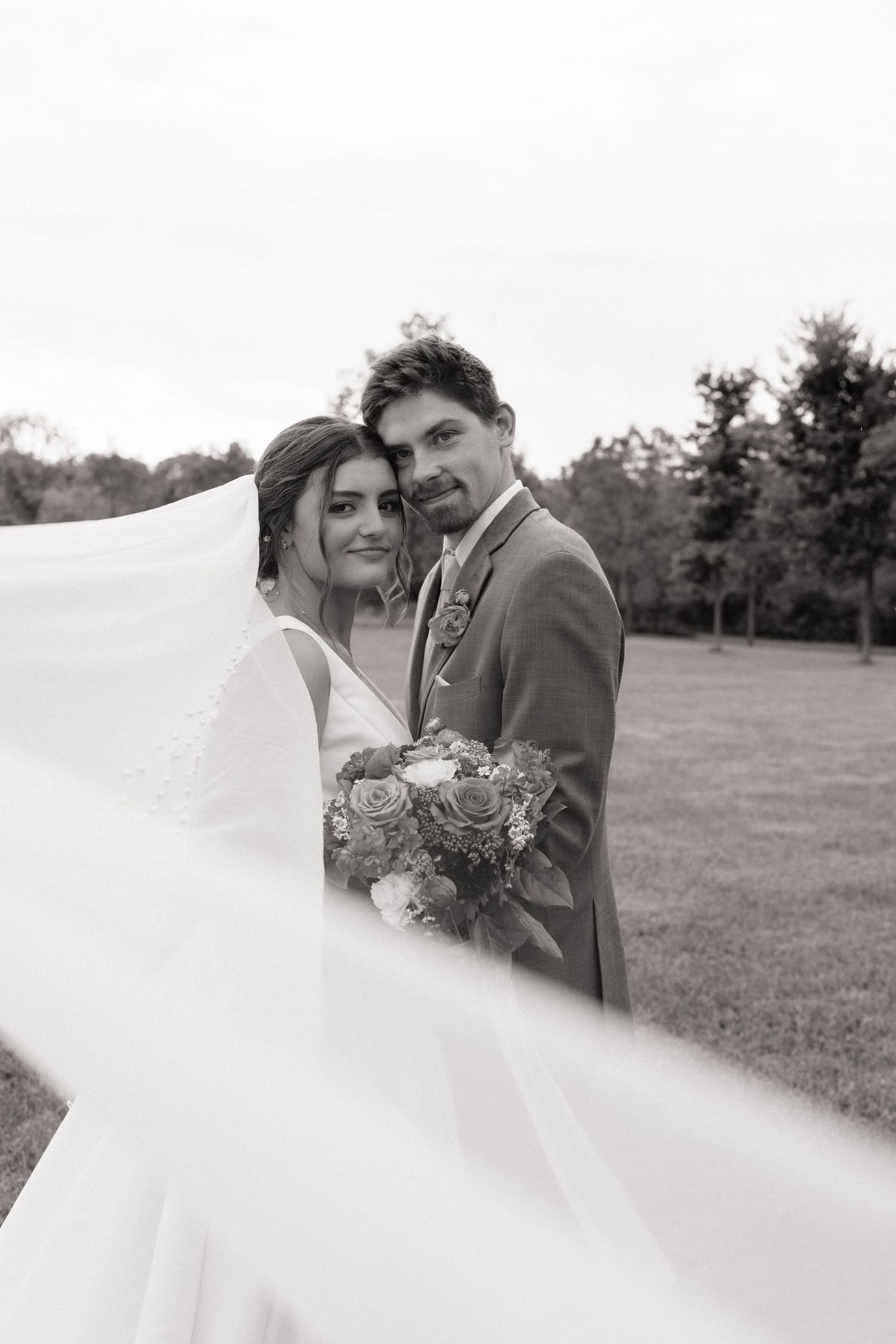 Black and white wedding photo of bride and groom embracing outdoors, veil flowing in wind, bride holding bouquet.