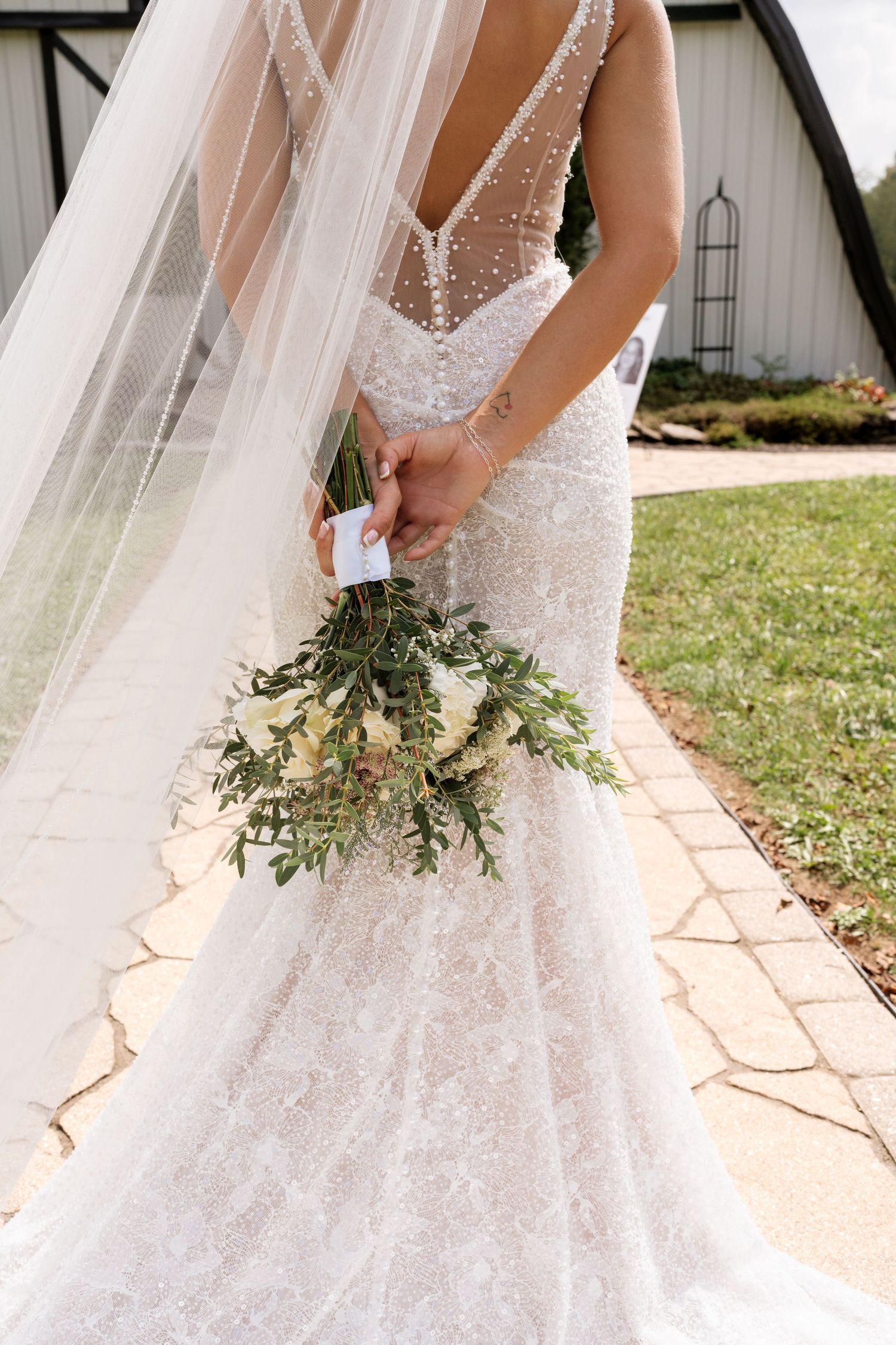 Bride in a pearl-embellished lace wedding gown holding a white and greenery bouquet, with a flowing veil, outdoors.
