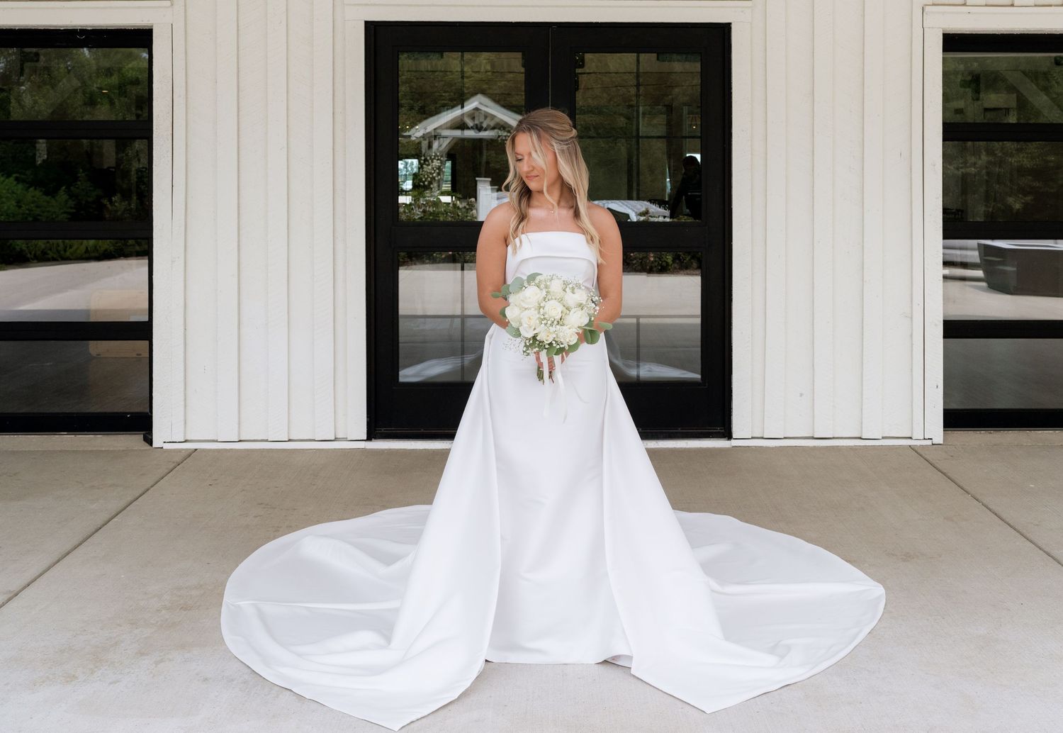Bride in strapless white ball gown with long train holding white bouquet standing before modern white building.