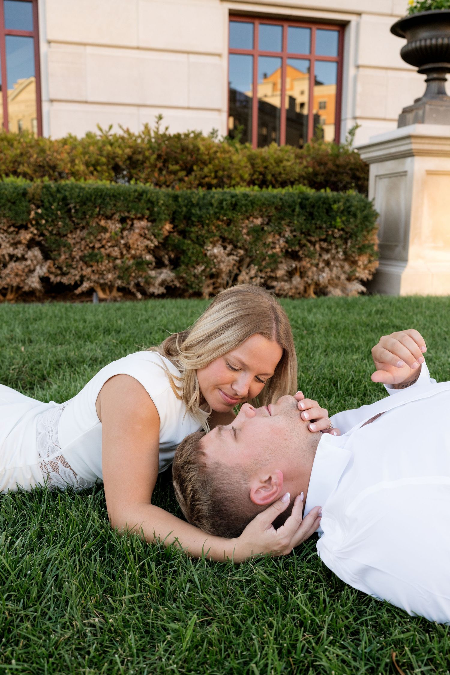 Couple in white outfits lying on green grass, woman tenderly holding mans face during romantic outdoor engagement photo.