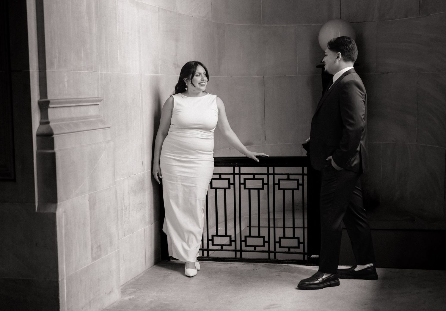 Black and white wedding photo of bride in white gown and groom in suit posing elegantly in a grand stone building.