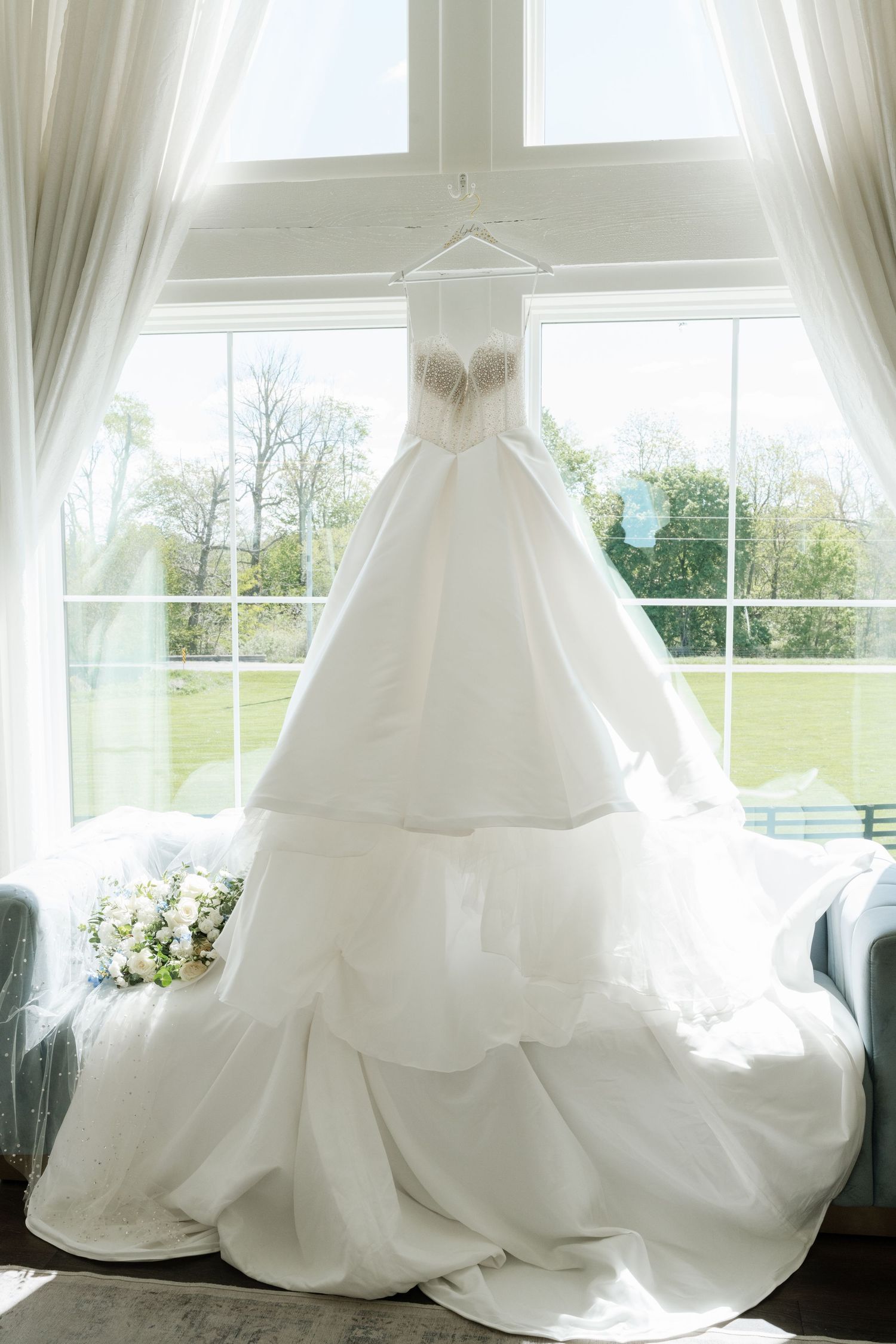 Elegant white strapless ball gown wedding dress hanging in window with tiered skirt and bridal bouquet on chair.