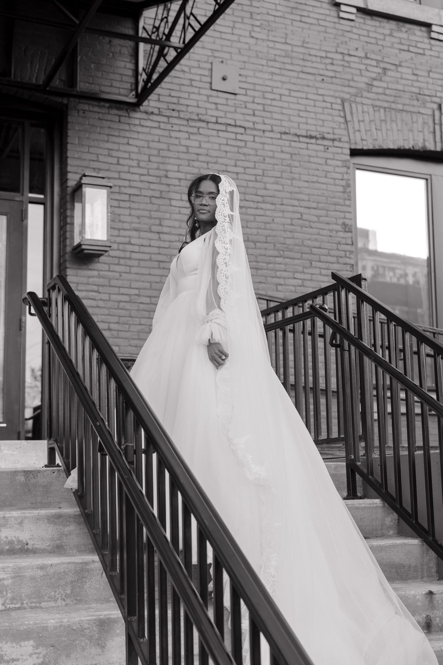 Bride in flowing white dress poses on brick building staircase with black railing.