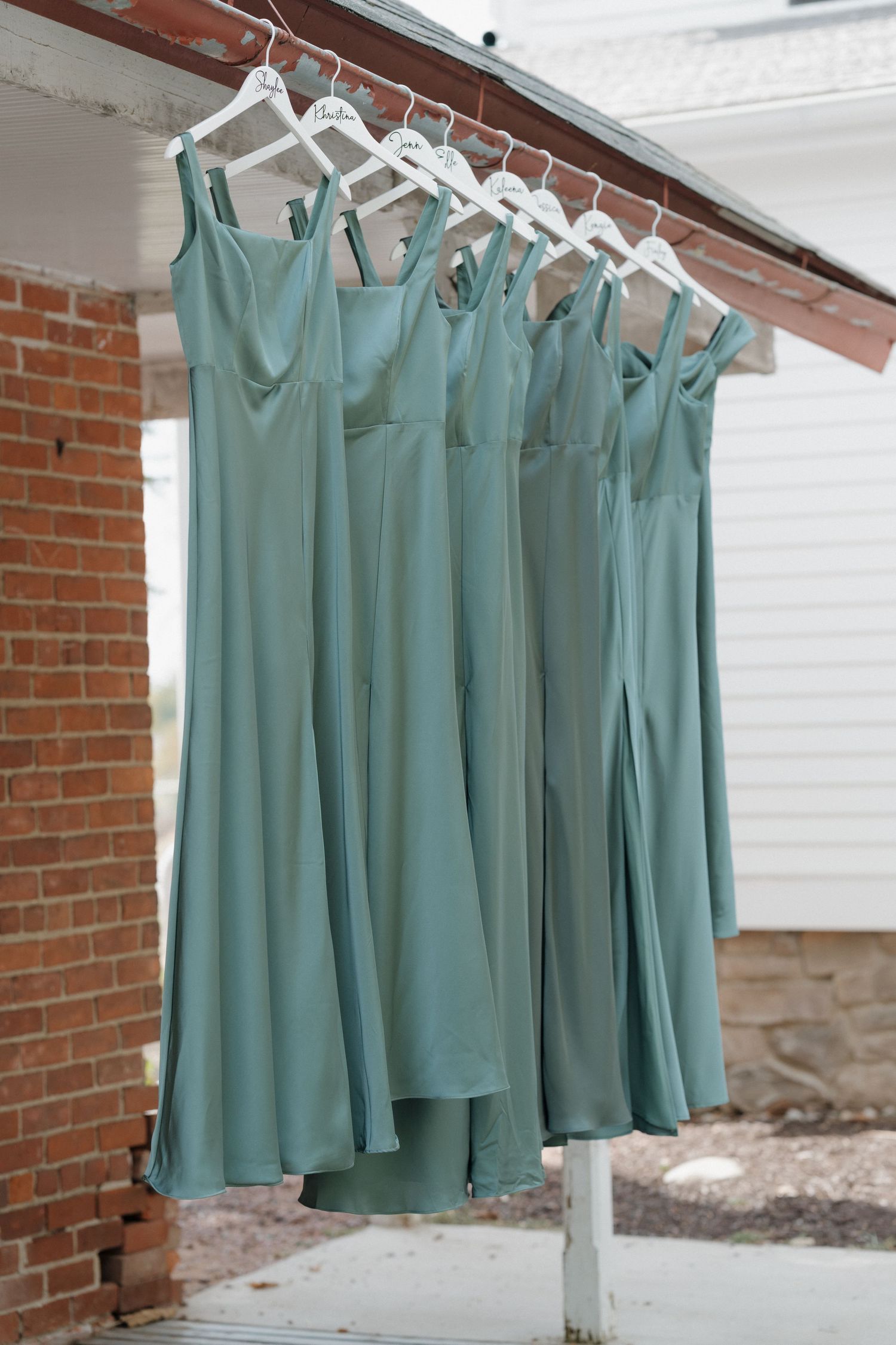 Row of matching sage green bridesmaid dresses hanging on white hangers against a brick wall.