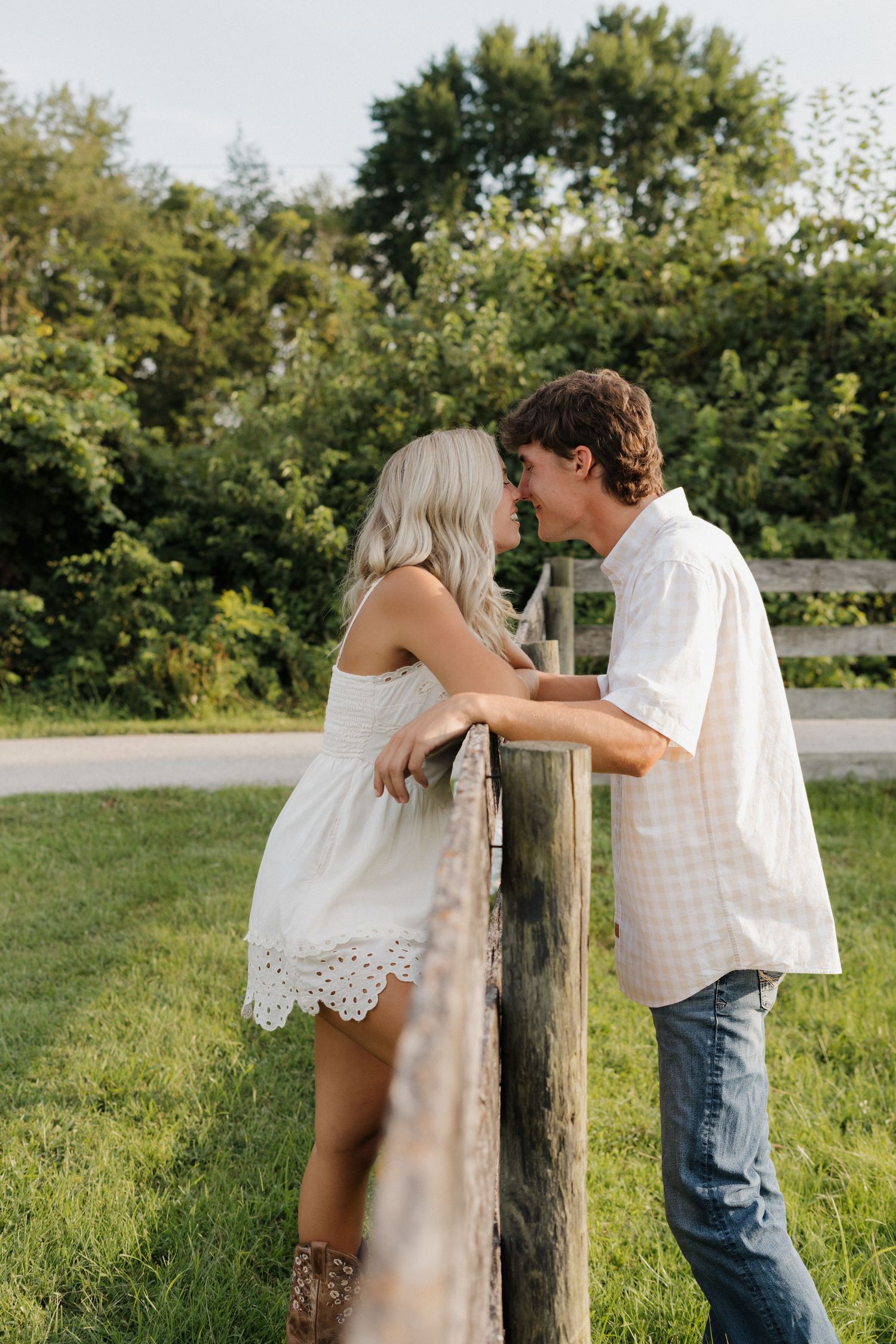 A couple shares a romantic moment leaning against a wooden fence post in golden sunlight.