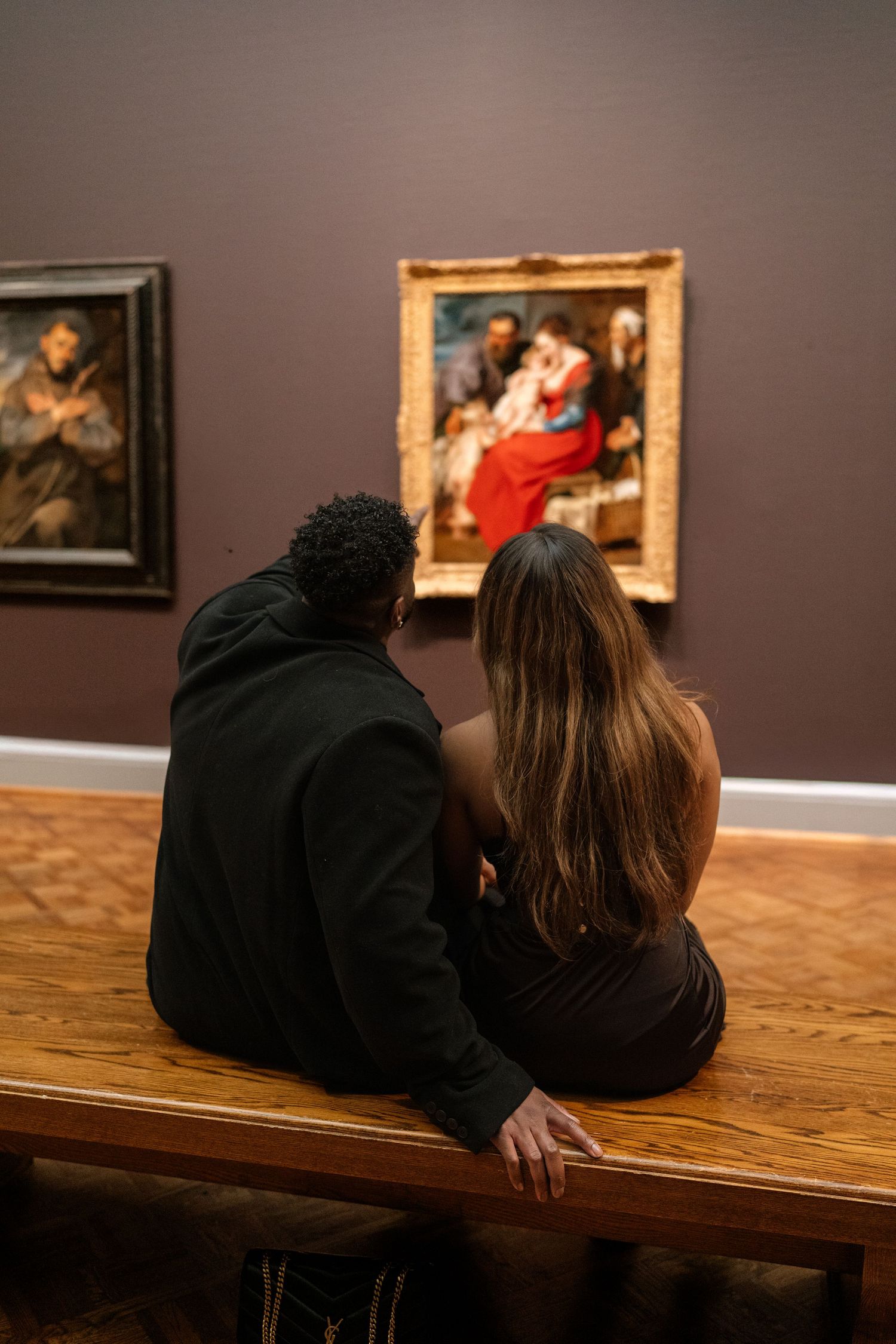 Two museum visitors lean close together while viewing artwork in a gallery setting.