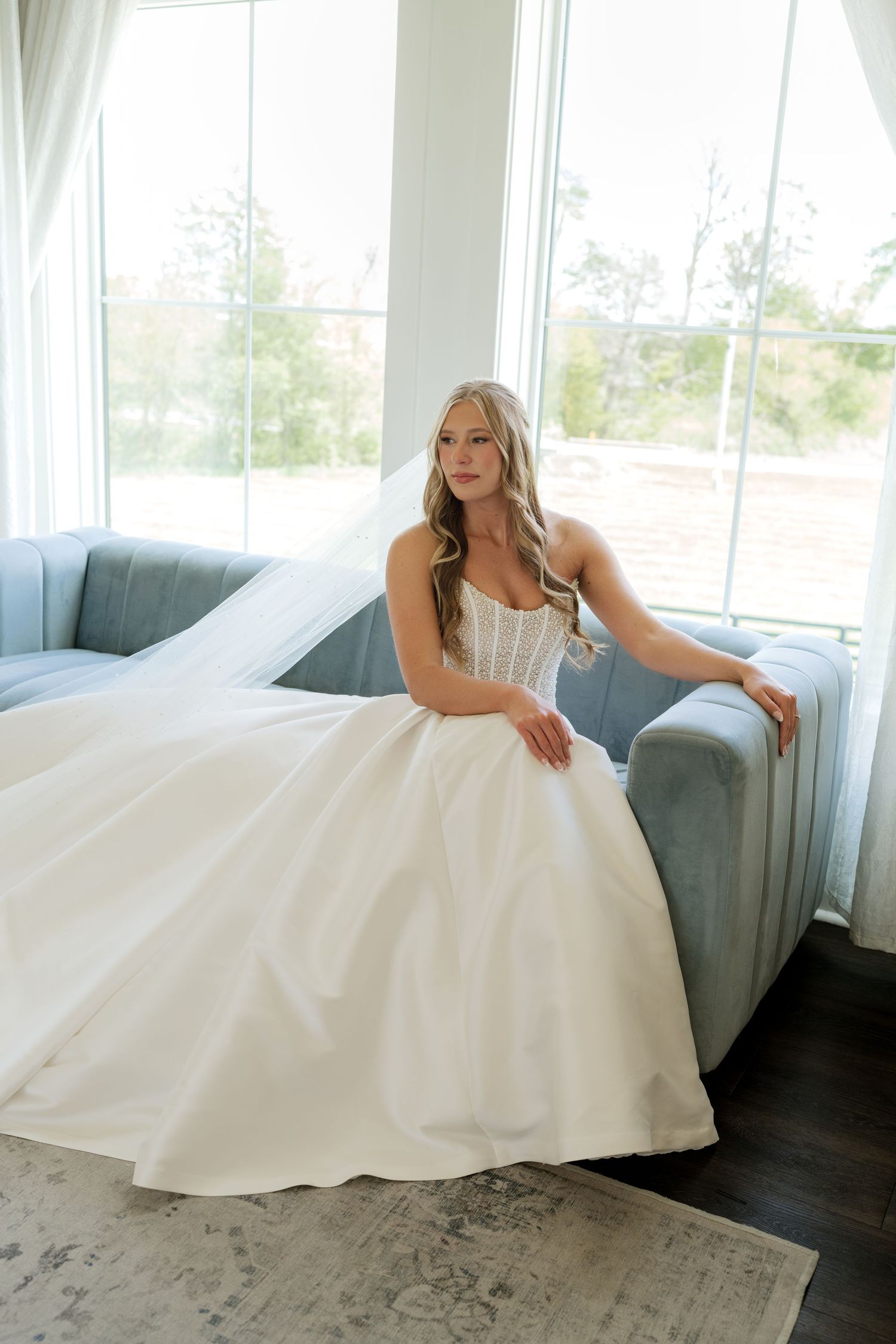 A bride in a white ballgown and veil sits gracefully on a blue sofa near large windows in a bright, airy room.