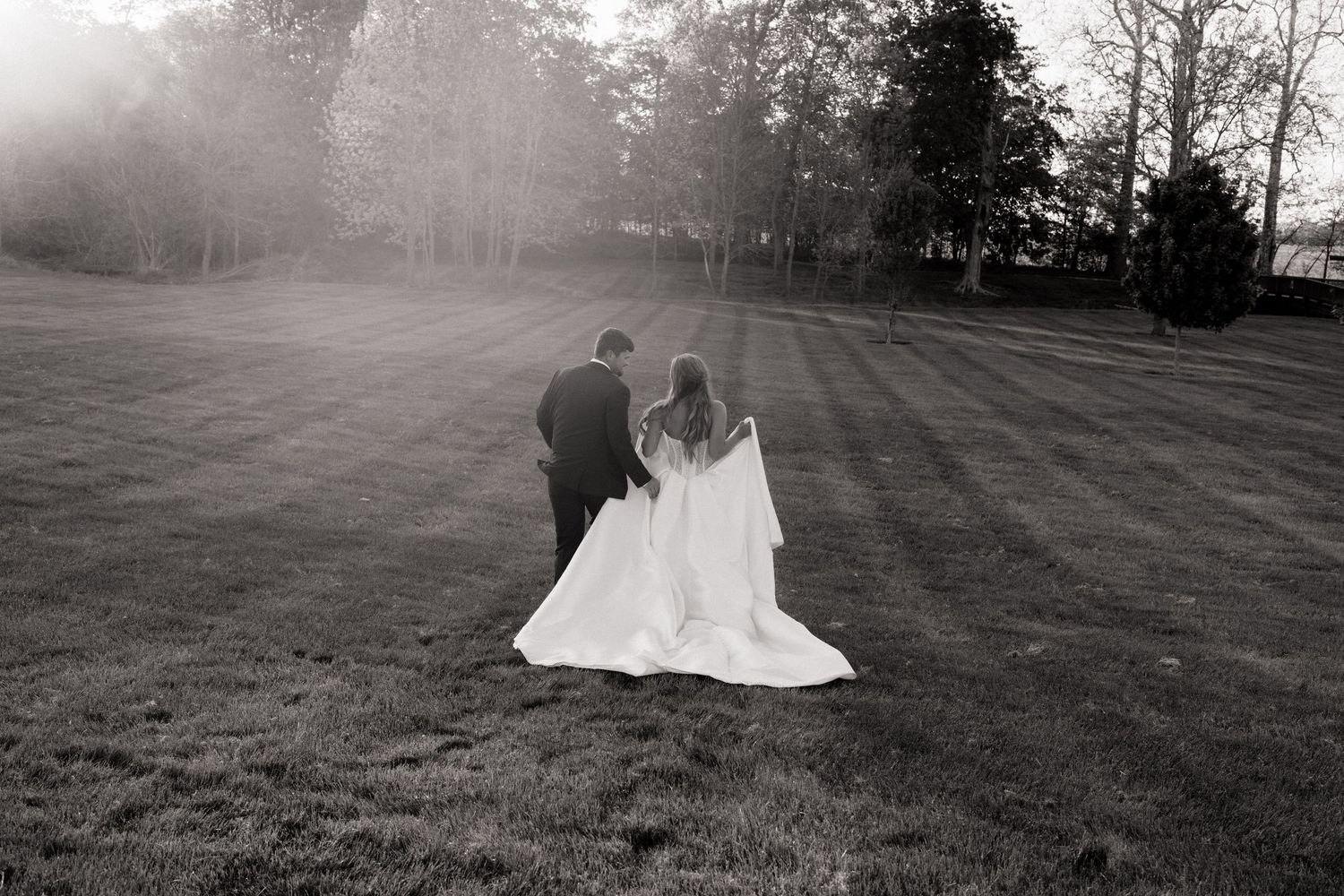 Black and white photo of bride and groom walking hand in hand across a lush lawn at sunset.
