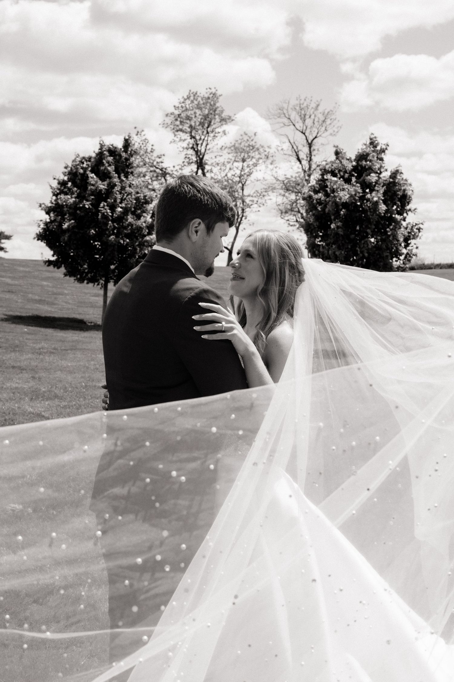 Black and white photo of a bride and groom nearly kissing outdoors, bride's veil flowing in the breeze.
