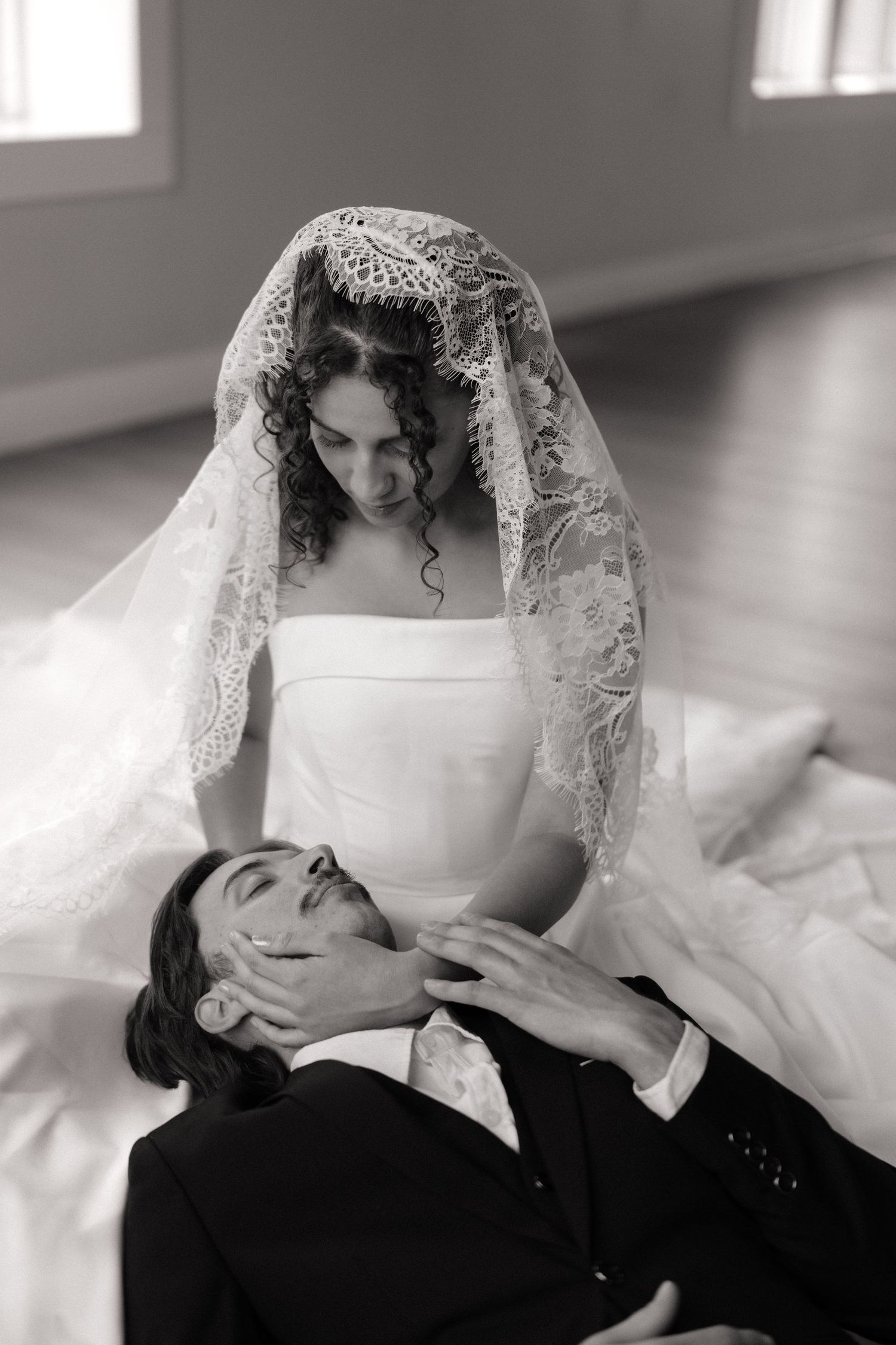 Black and white wedding photo of bride in lace veil cradling groom's face as he lies in her lap on floor.
