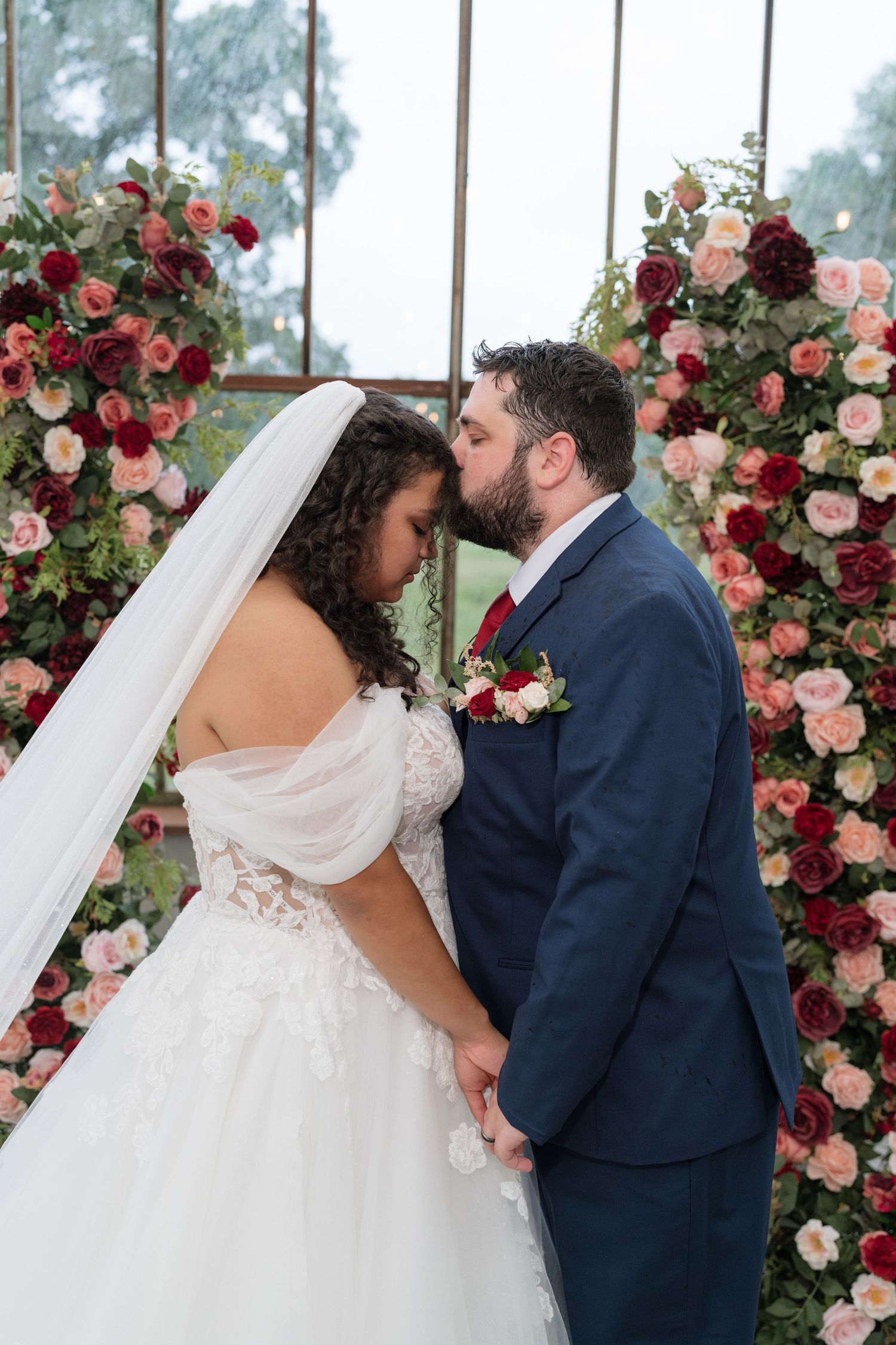 Bride and groom share first kiss at floral arch adorned with pink and red roses during romantic wedding ceremony.