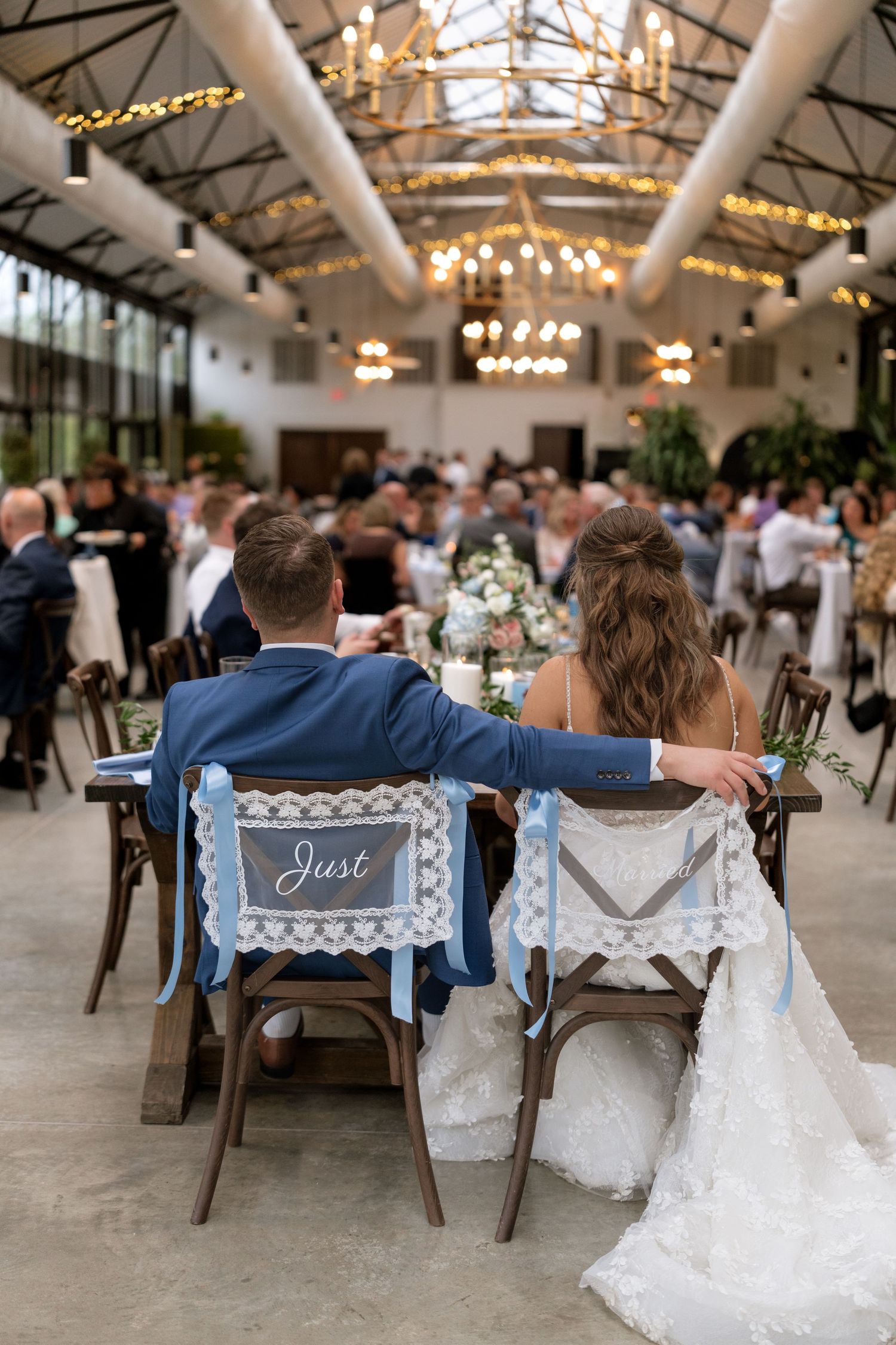 Bride and groom seated at sweetheart table with lace chair signs at elegant indoor wedding reception.