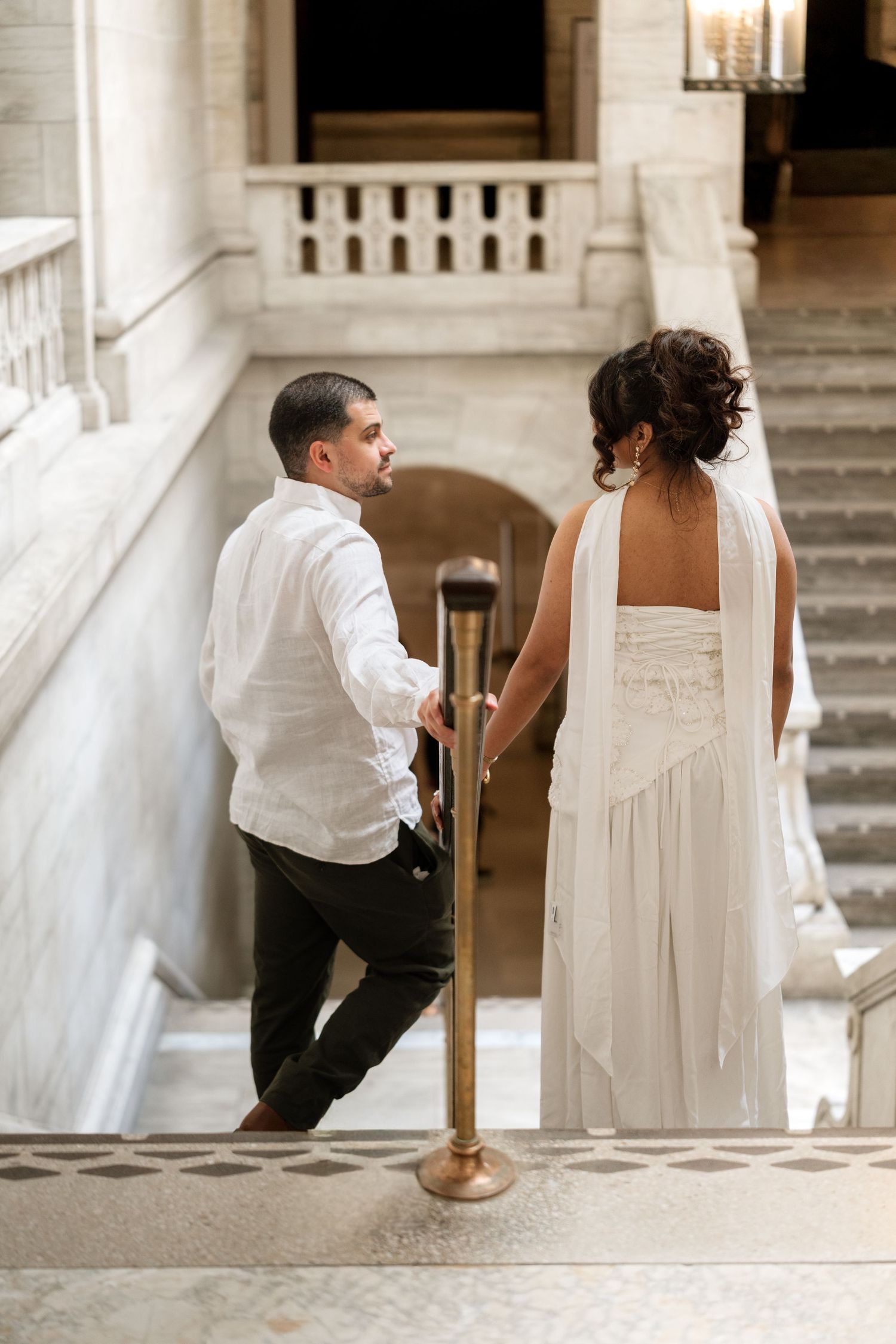 Bride in white gown and groom in white shirt hold hands ascending marble staircase in elegant venue.