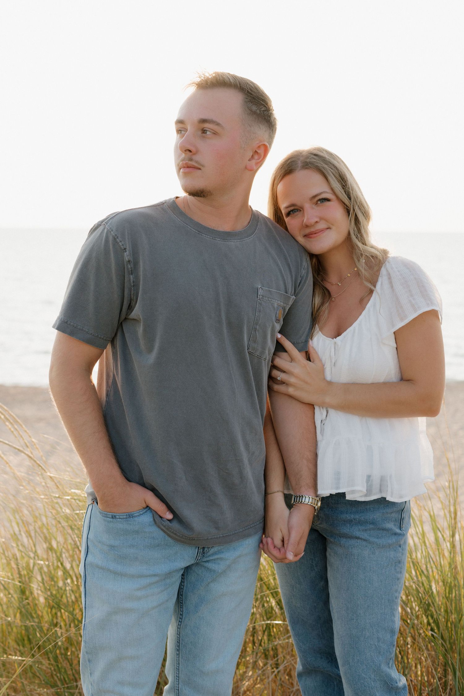 A couple poses together on a beach, woman in white top leans on man in gray tee, both wearing jeans.