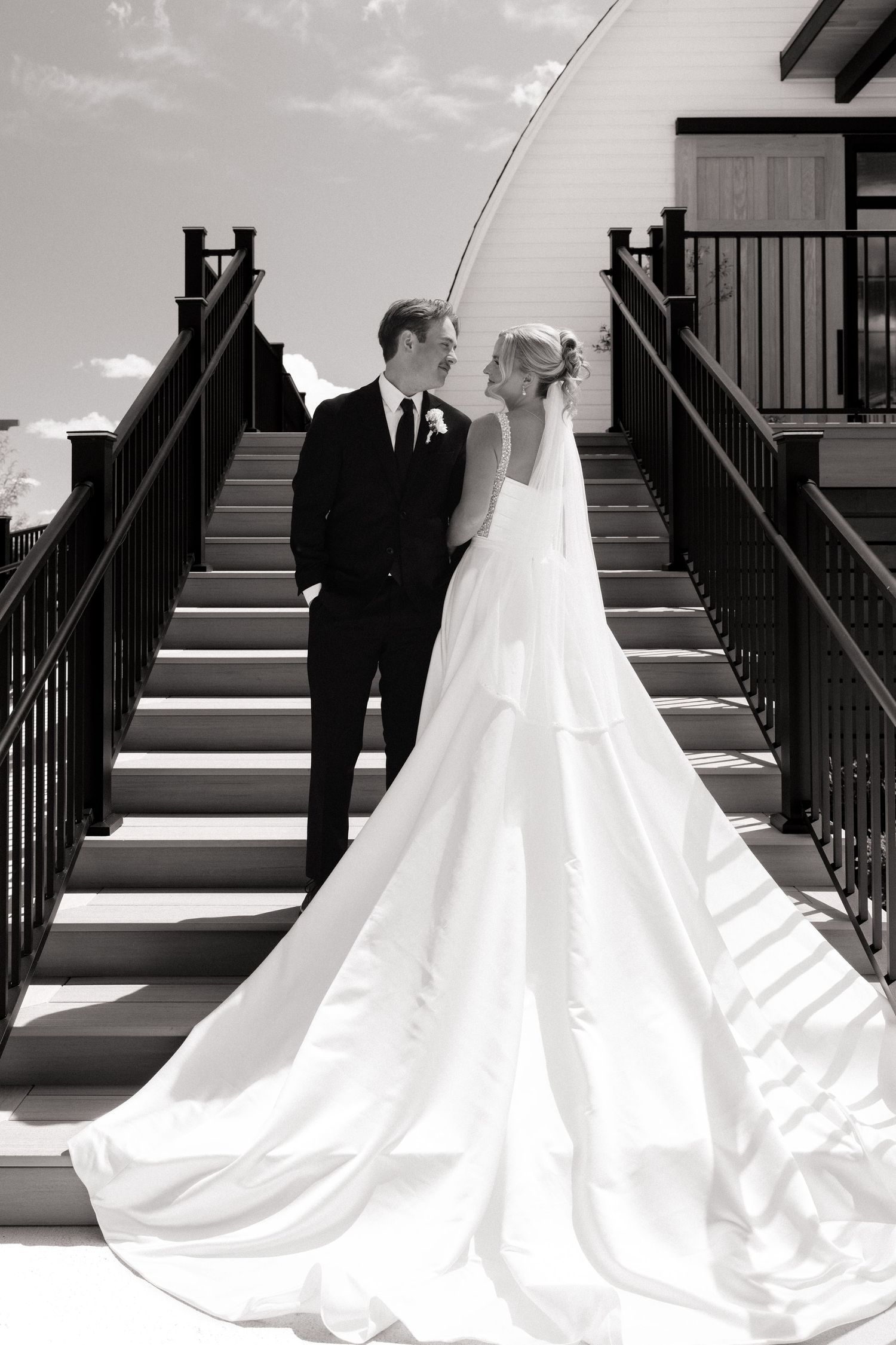Black and white photo of bride and groom standing on outdoor staircase, bride wearing elegant gown with long train.