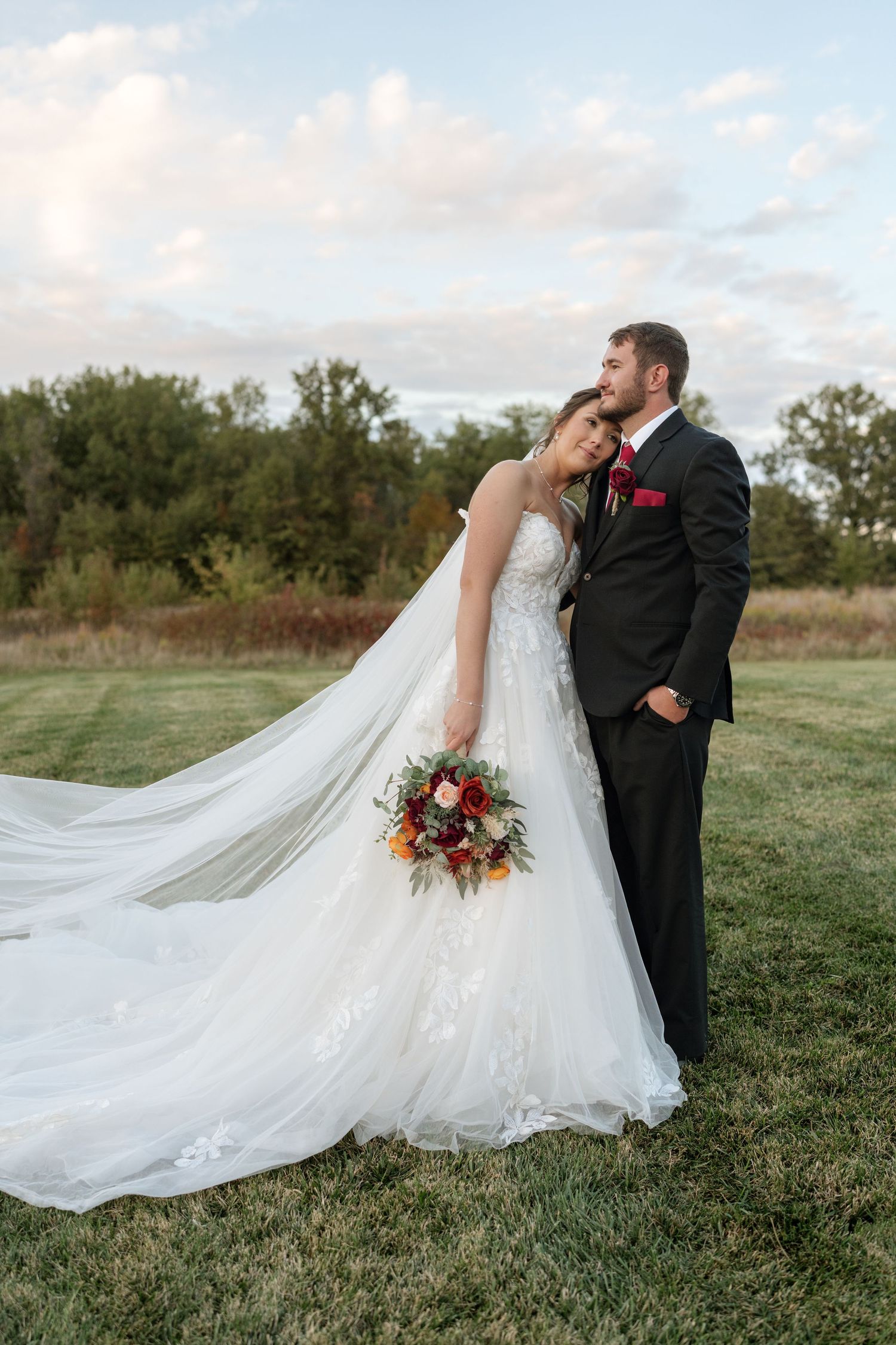 Romantic wedding portrait with flowing veil and red bouquet against natural landscape.