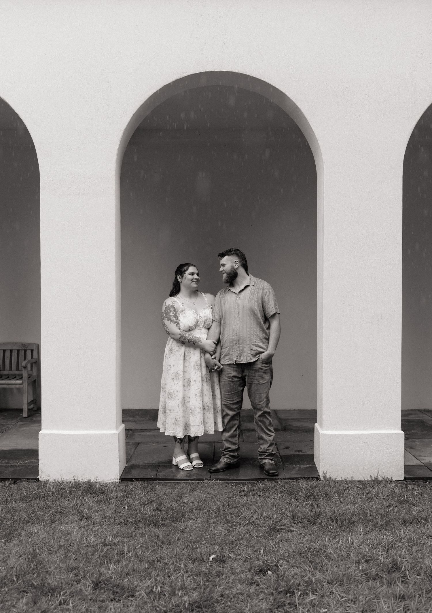 Black and white photo of a couple holding hands under white arched colonnade, woman in floral dress, man in casual attire.
