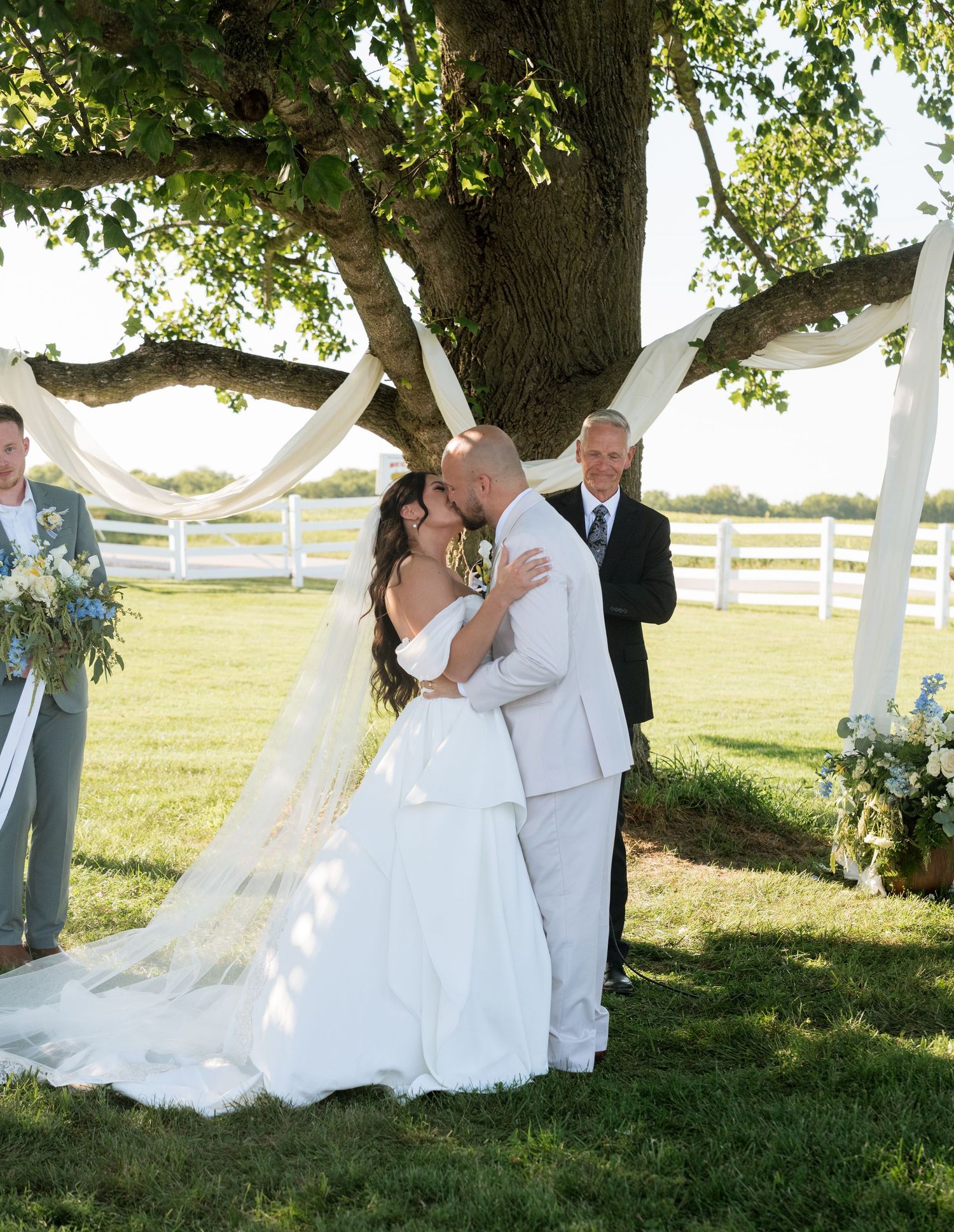 Bride and groom share first kiss at outdoor wedding ceremony under large tree with white fabric draping.