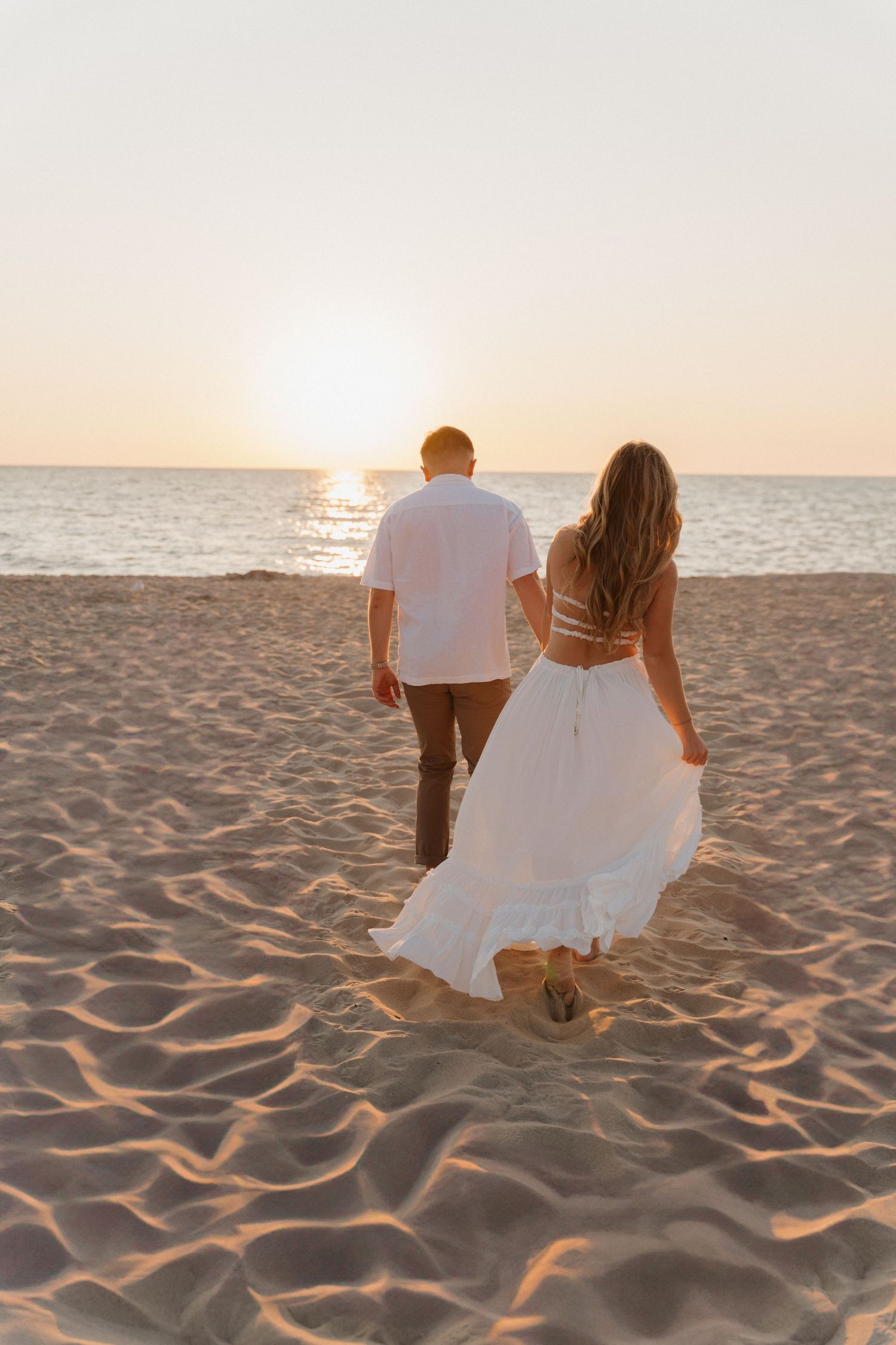 A couple in white clothing sits together on a sandy beach watching the sunset over the ocean.