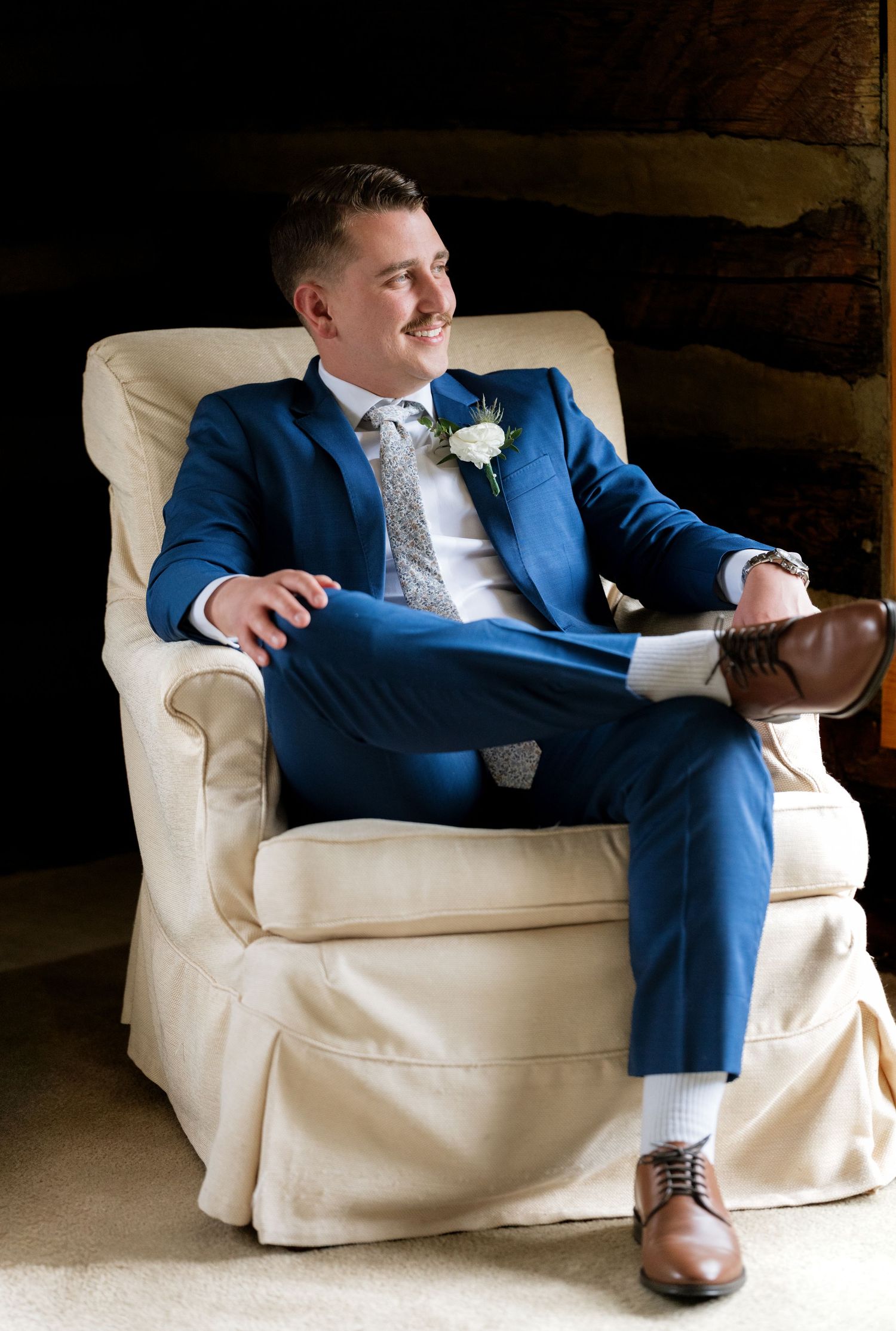 A groom in a blue suit with boutonniere sits relaxed in a cream armchair, smiling, wearing brown dress shoes.