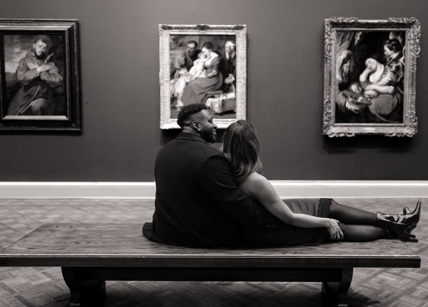 Couple sitting on museum bench viewing classic paintings in black and white art gallery setting.