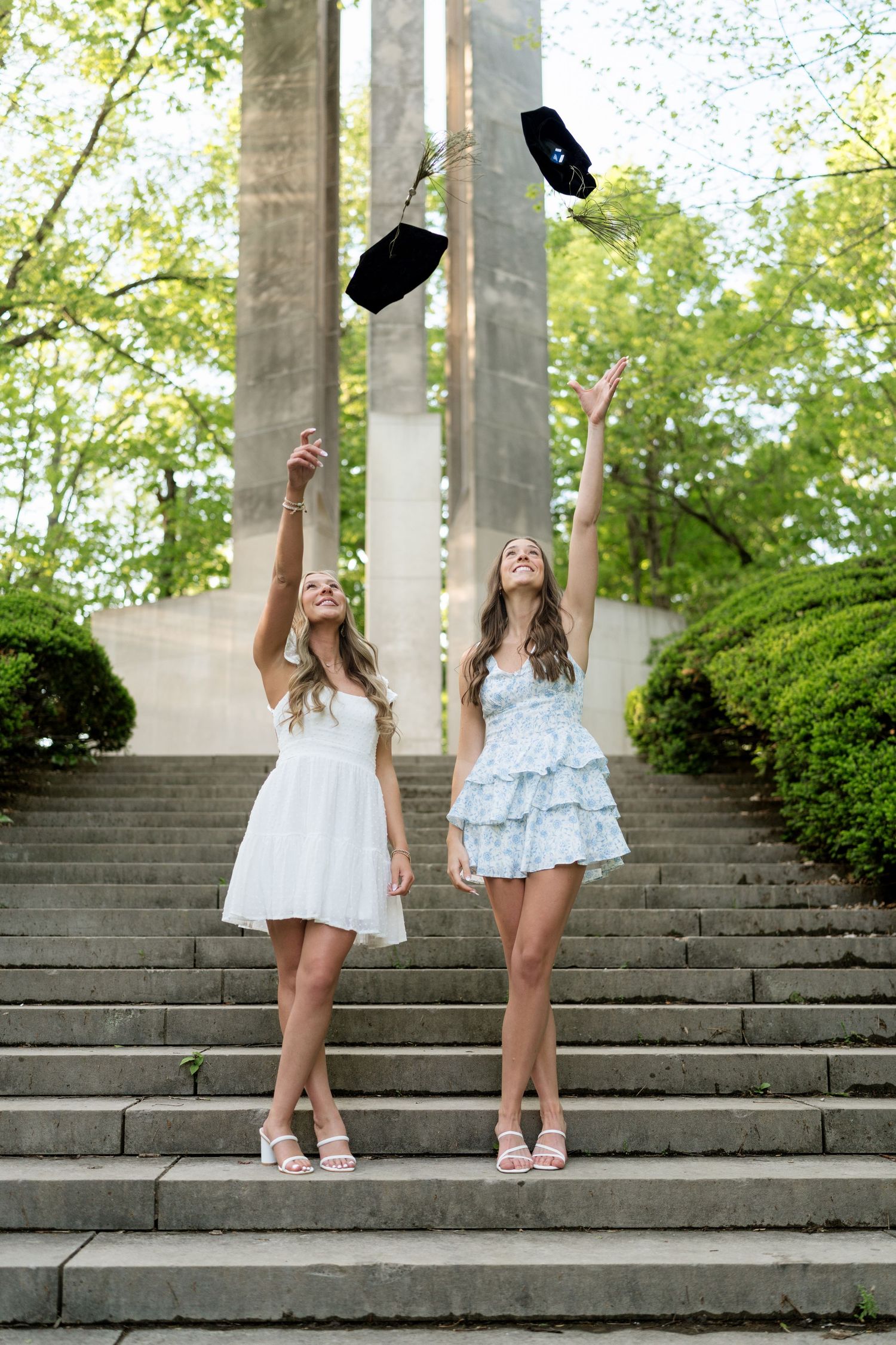 Two young women in white dresses tossing graduation caps on stone steps outdoors.