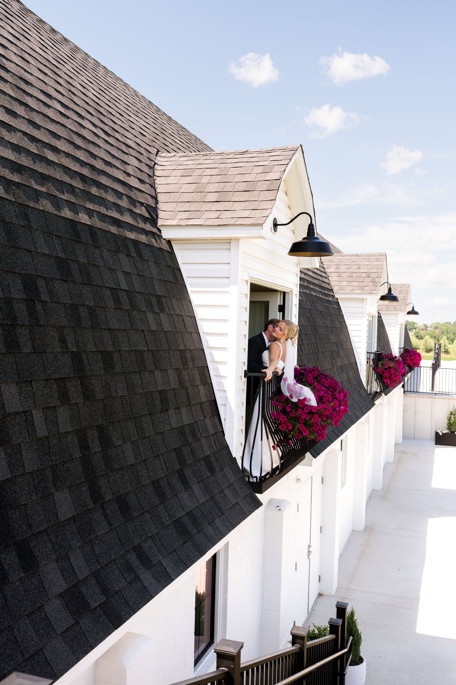 Couple sharing a romantic kiss on a charming white balcony adorned with pink flowers beneath dark shingled rooftops.