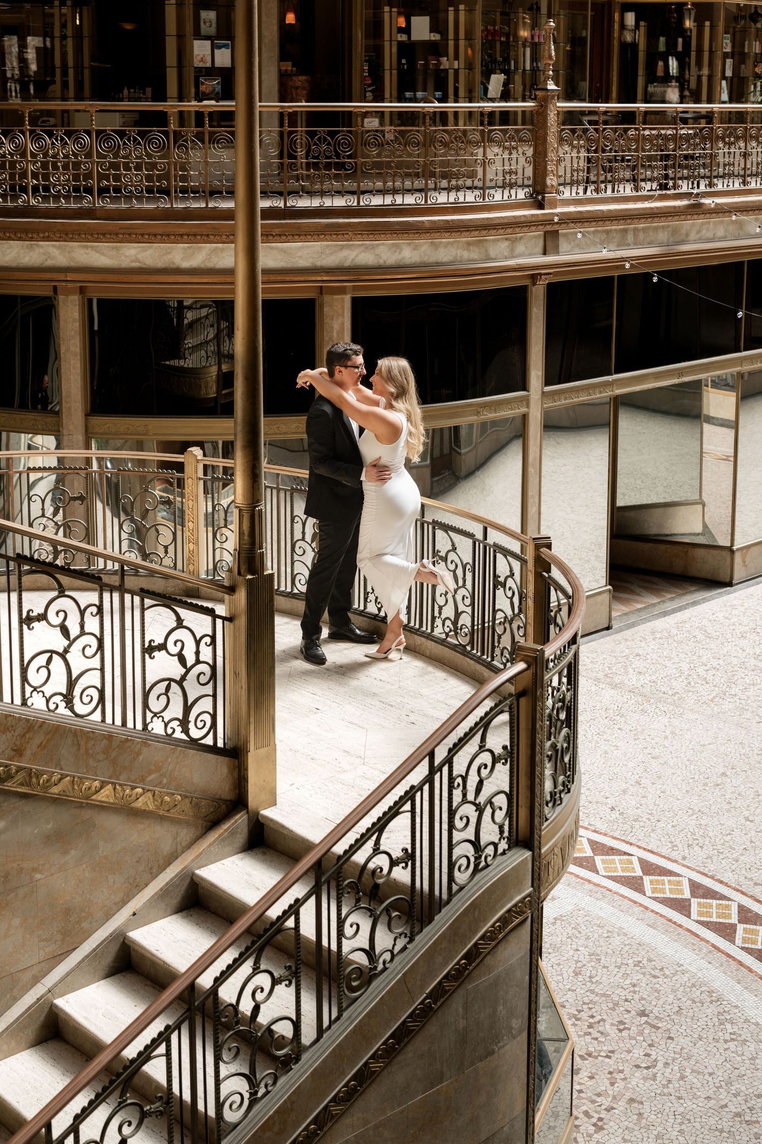 Couple embracing on ornate iron balcony of elegant historic building with decorative spiral staircase.