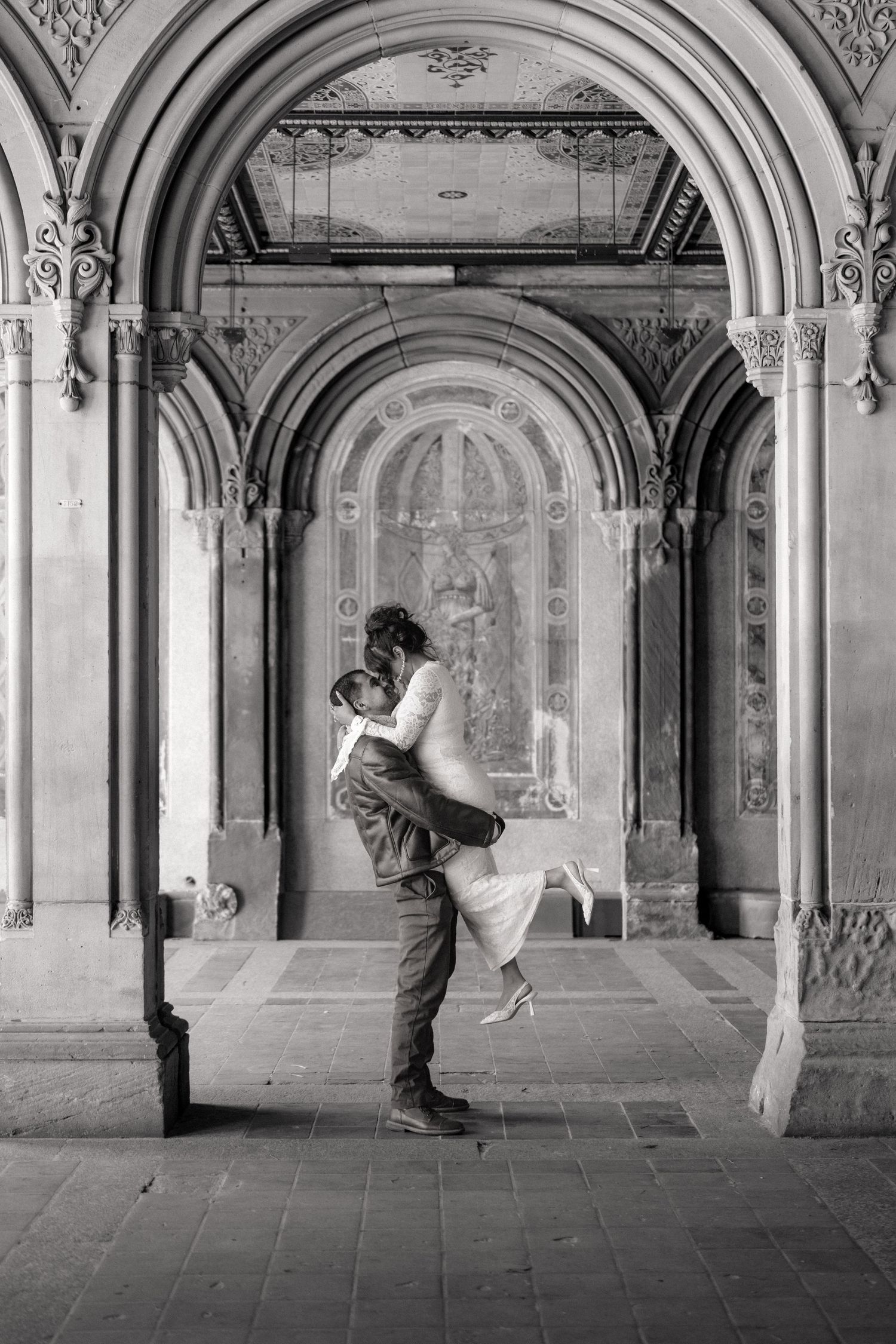 Black and white photo of a couple embracing under ornate stone arches at Bethesda Terrace, Central Park.