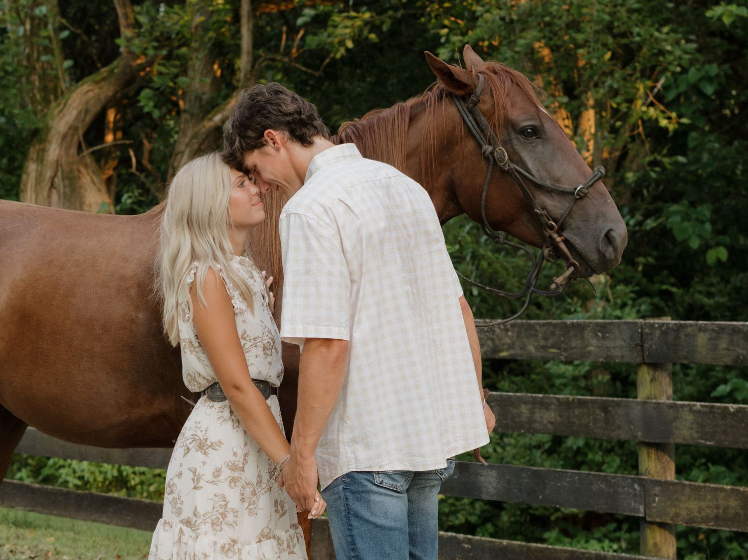 A couple stands with their horse near a wooden fence in a lush garden setting.