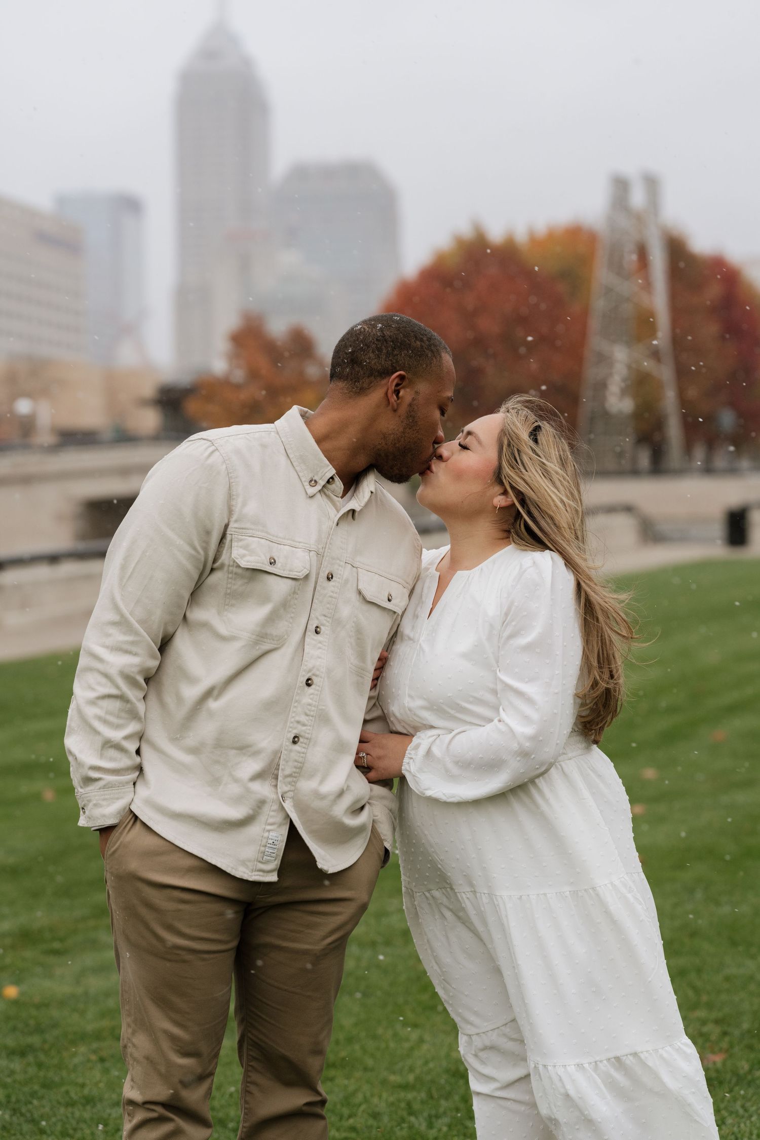 A couple shares romantic moments together on a grassy hill with an urban skyline in the background during autumn.