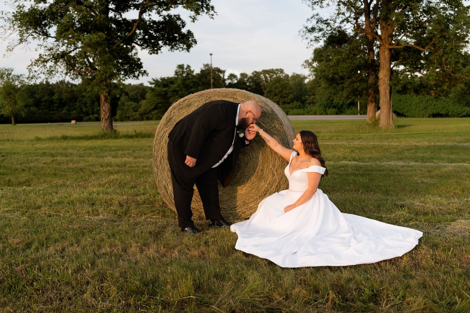 Bride in white gown sits by hay bale as groom in tuxedo leans in romantically in a green field.