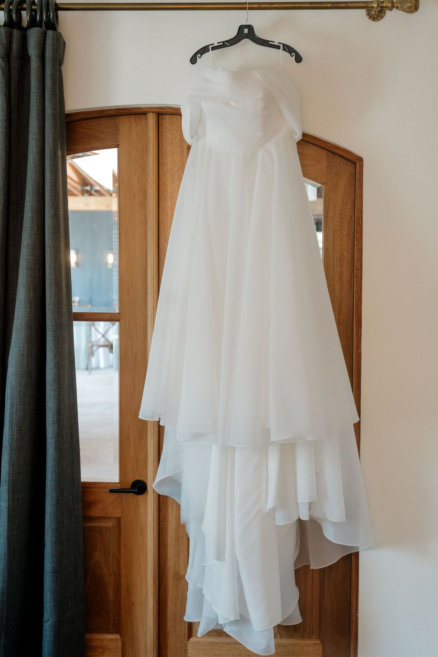 White wedding dress hanging on display against wooden doors and dark curtains.