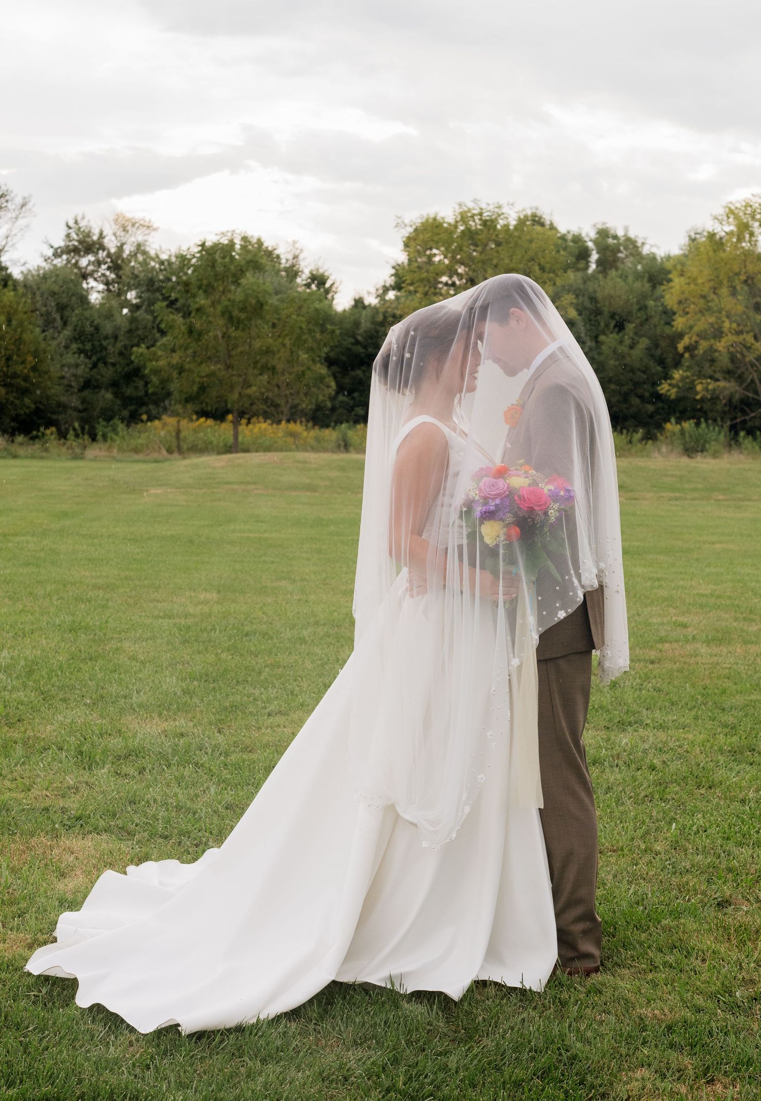 Bride and groom kissing under a flowing white veil in a green field on their wedding day.