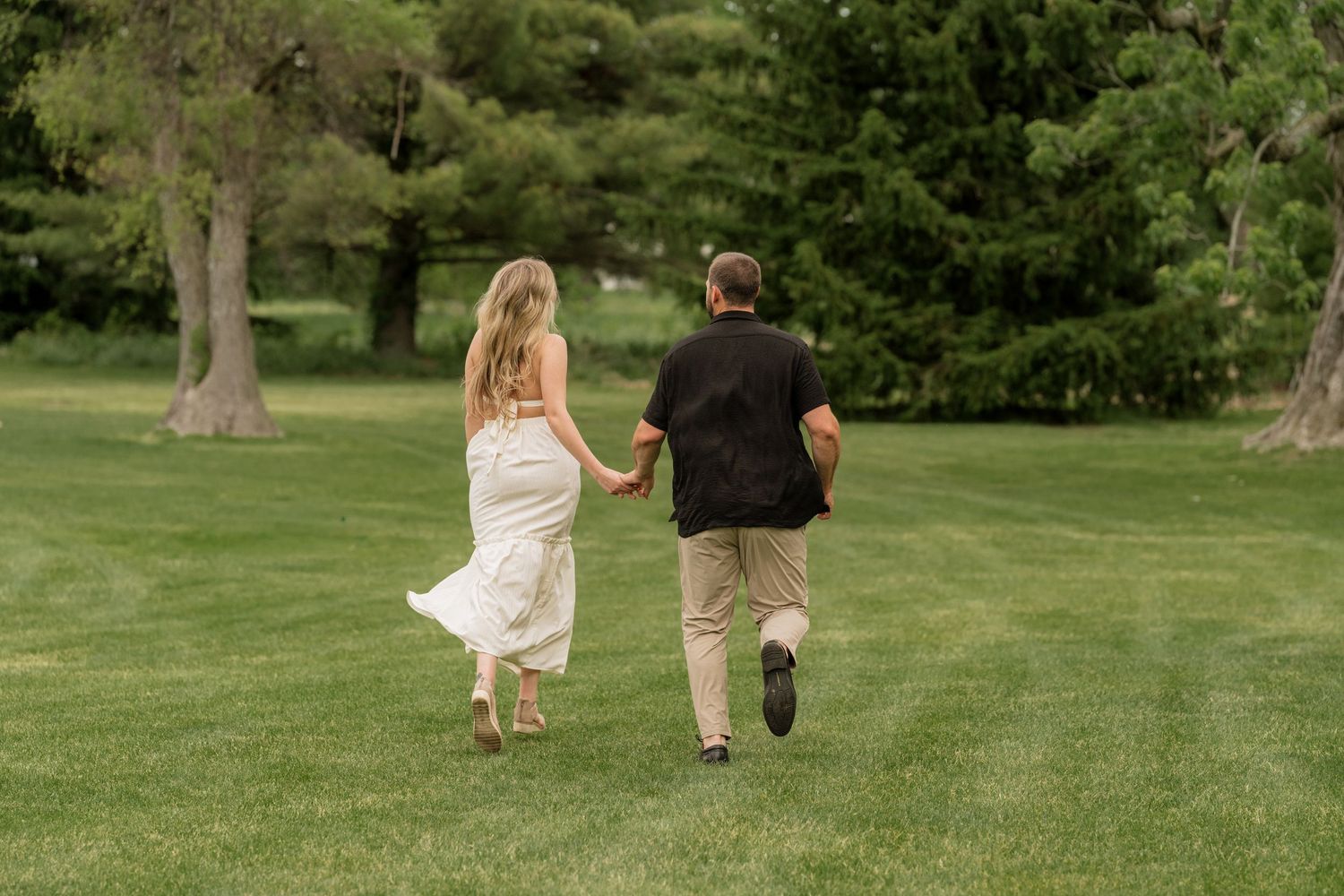 Couple holding hands running across lush green lawn, woman in white dress, man in black shirt, trees in background.
