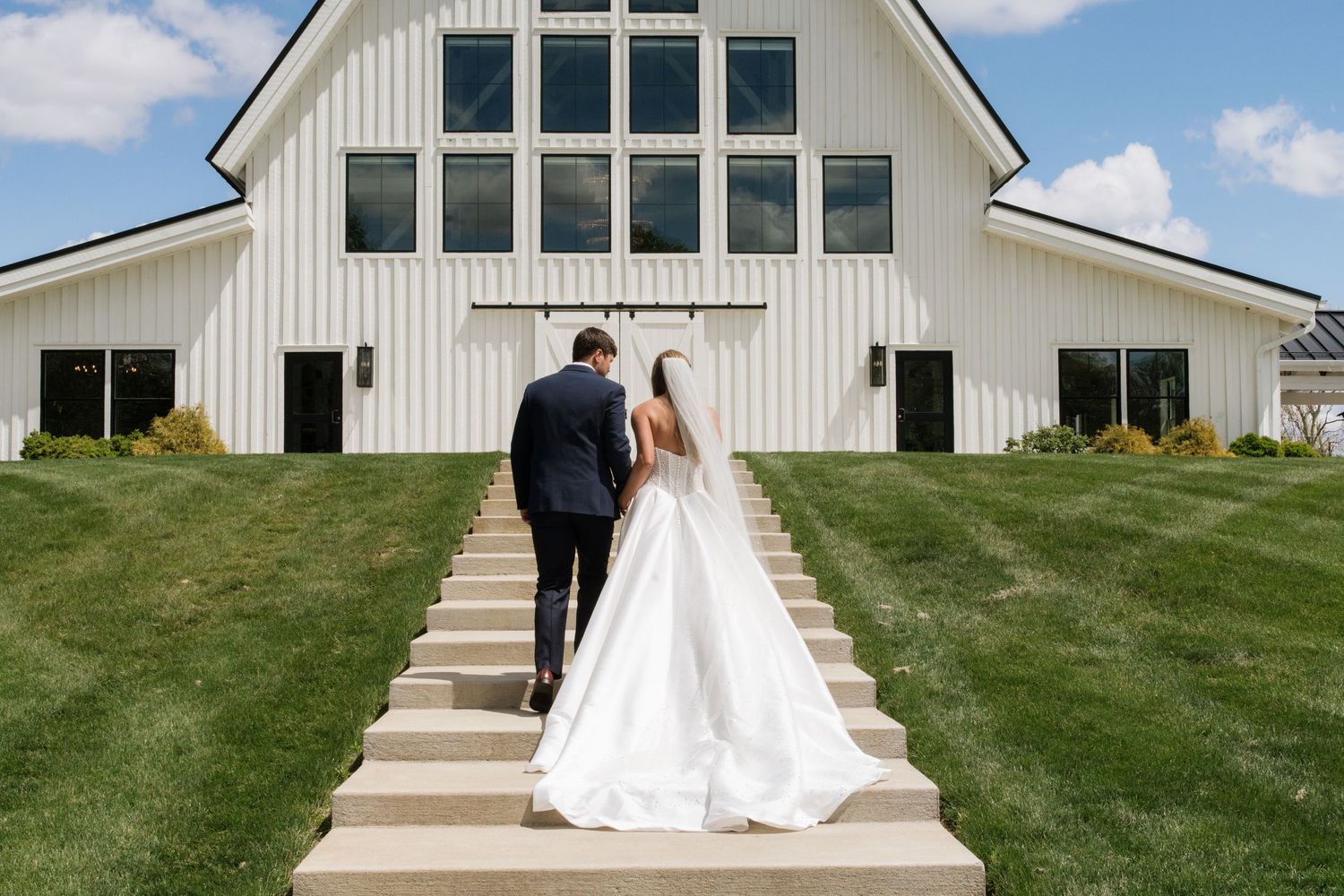 Bride and groom walk up stone steps toward a white barn wedding venue surrounded by lush green grass.