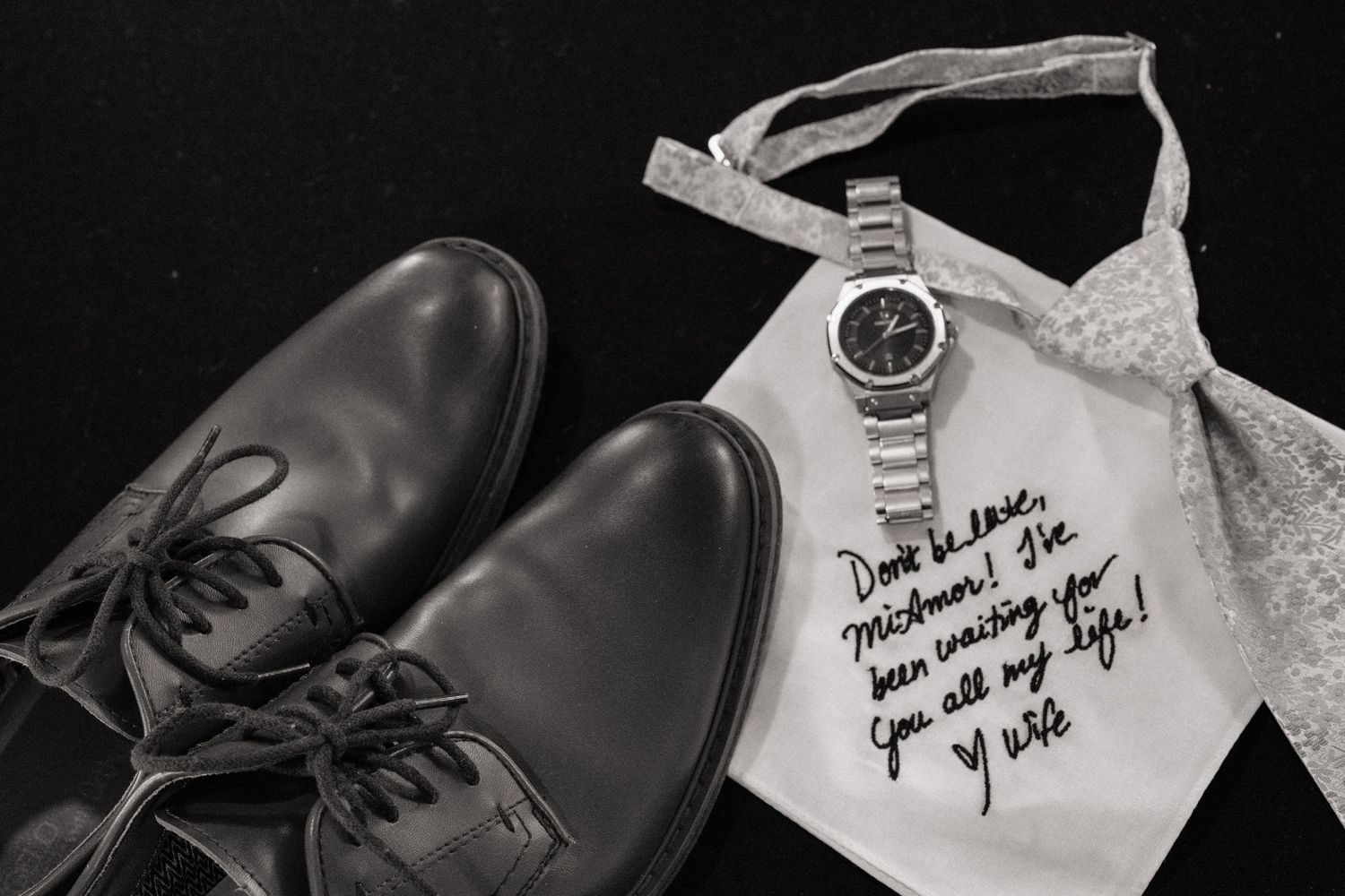 Black dress shoes, silver watch, and a handwritten note on white fabric with a bow tie on dark background.