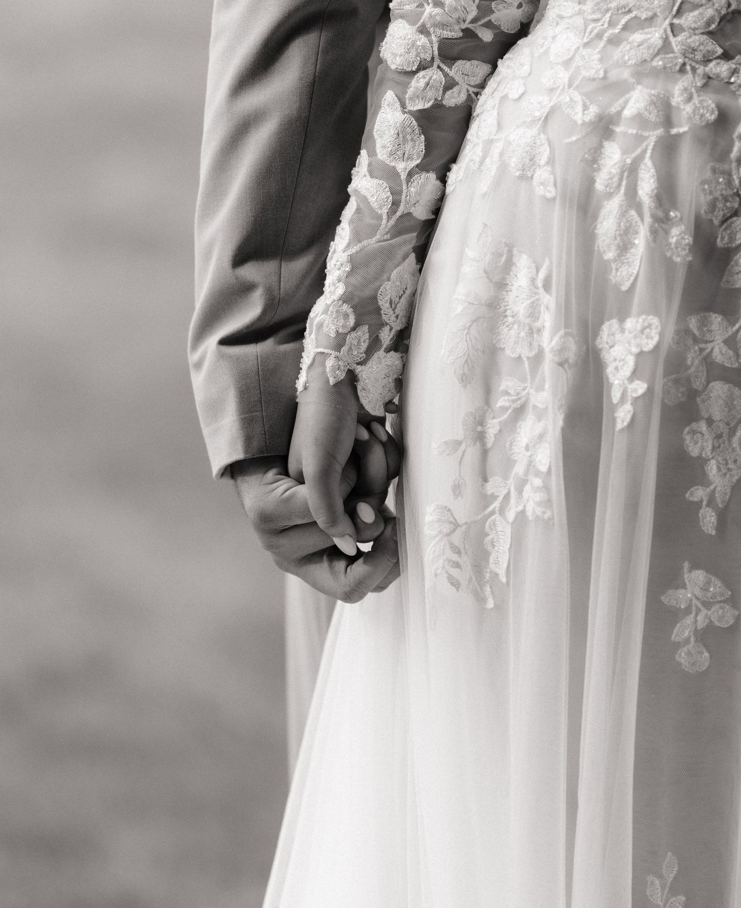 Black and white photo of a bride and groom holding hands, showcasing a floral lace wedding gown.