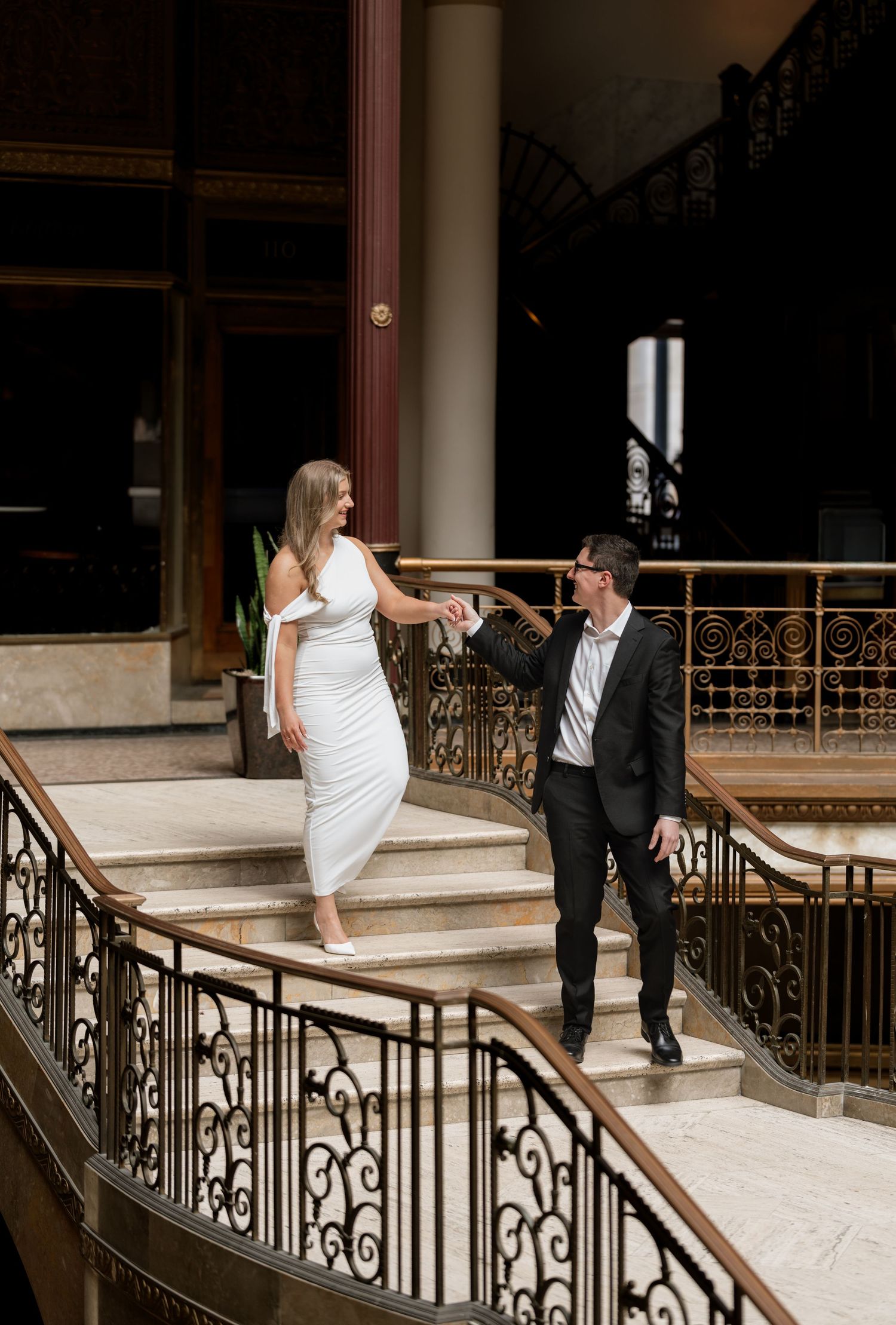 Couple in elegant attire holding hands on ornate staircase, woman in white dress, man in dark suit, grand interior.