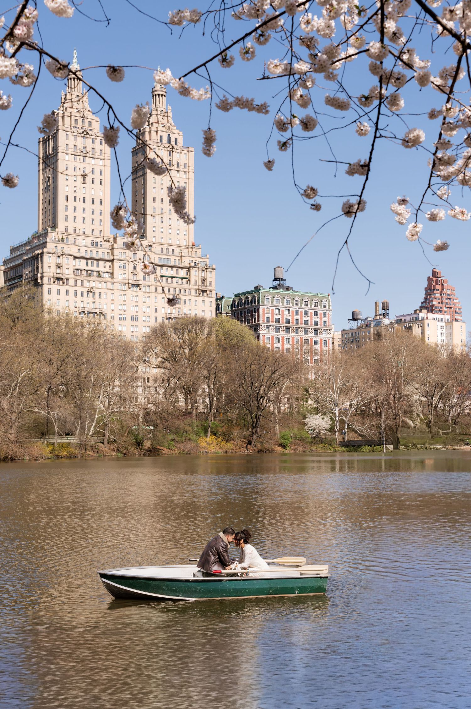 Couple in a rowboat on a serene Central Park lake with cherry blossoms and iconic NYC buildings in spring.