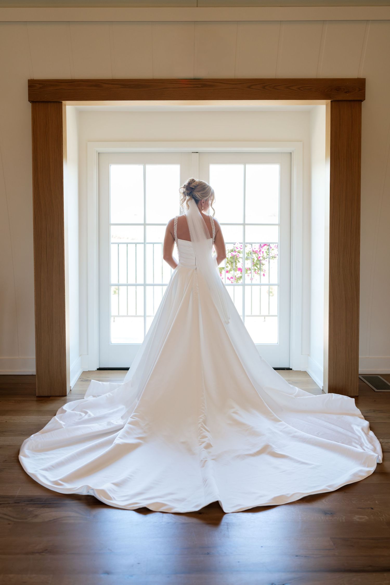 Bride in elegant white wedding gown with long train stands before French doors holding pink bouquet.