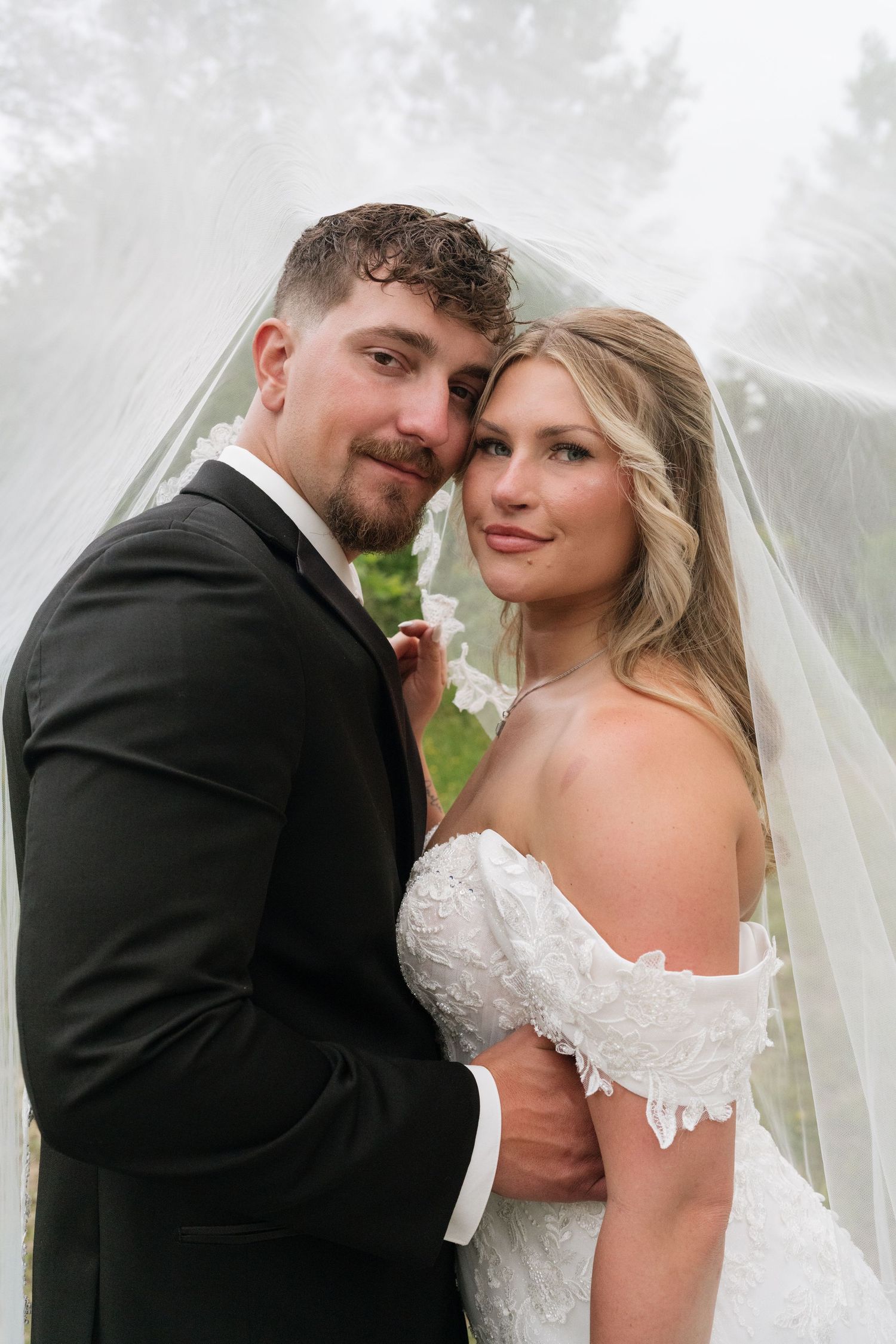 A bride and groom pose romantically under a flowing white veil outdoors on their wedding day.