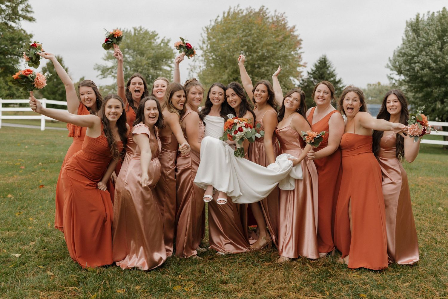 Wedding party in rust and blush colored dresses poses together on autumn grass.