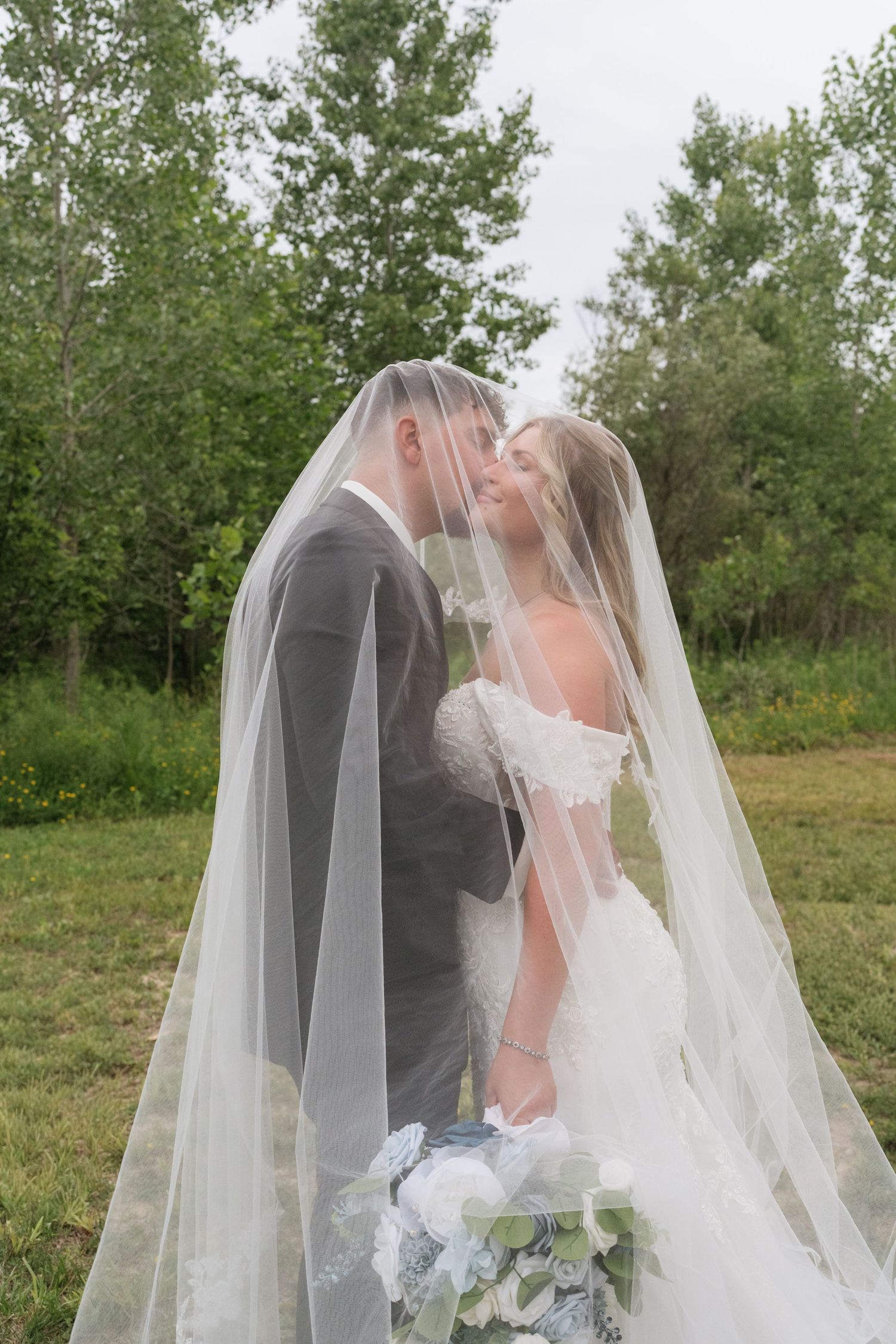 Bride and groom sharing a romantic moment under a flowing wedding veil outdoors, surrounded by lush green trees.